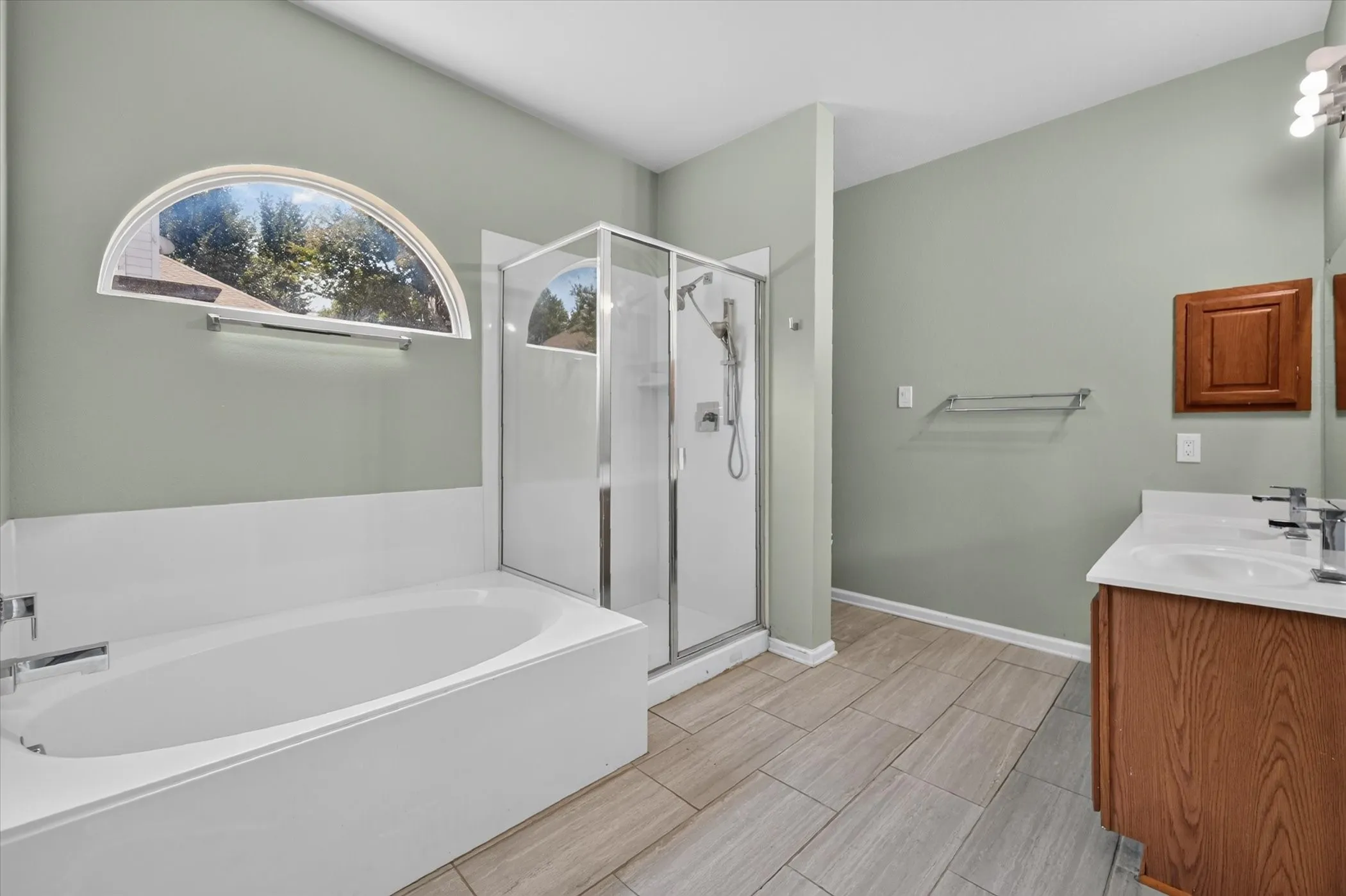 Alcove bathtub with an arched window, glass-enclosed shower, dual vanity with undermount sinks, and tile flooring