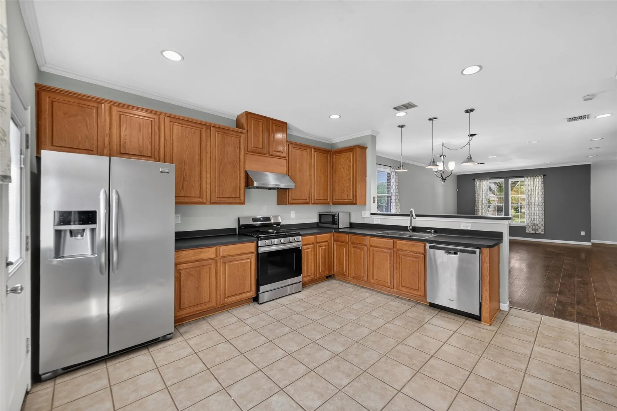 Spacious kitchen featuring warm wood cabinetry, stainless steel appliances including a side-by-side refrigerator and gas range, black countertops, tile flooring, recessed and pendant lighting, and a large island with seating area