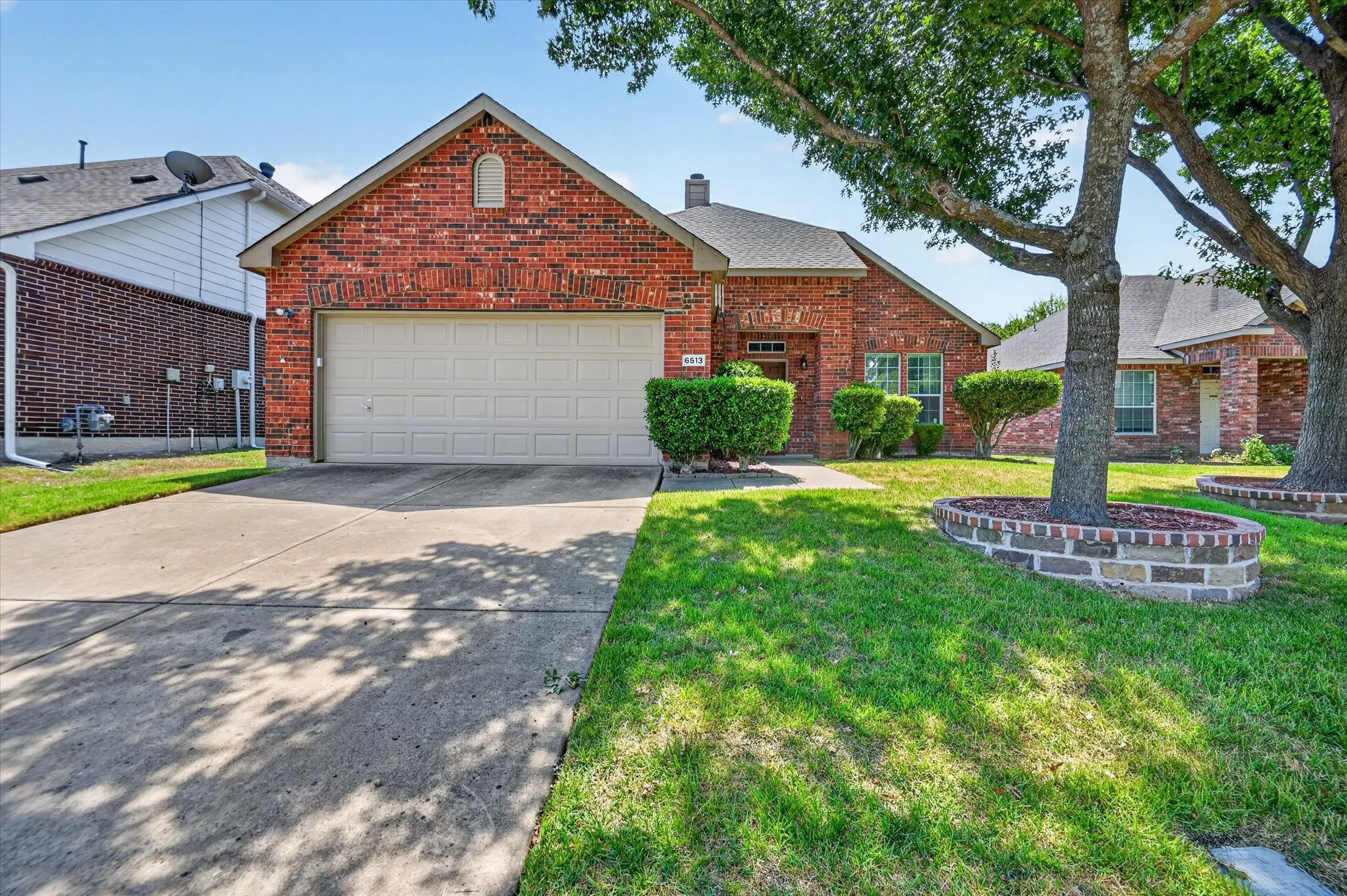 Charming brick home featuring a two-car garage, manicured front lawn with mature landscaping, brick front planter bed, and tree-lined lot with established shade trees