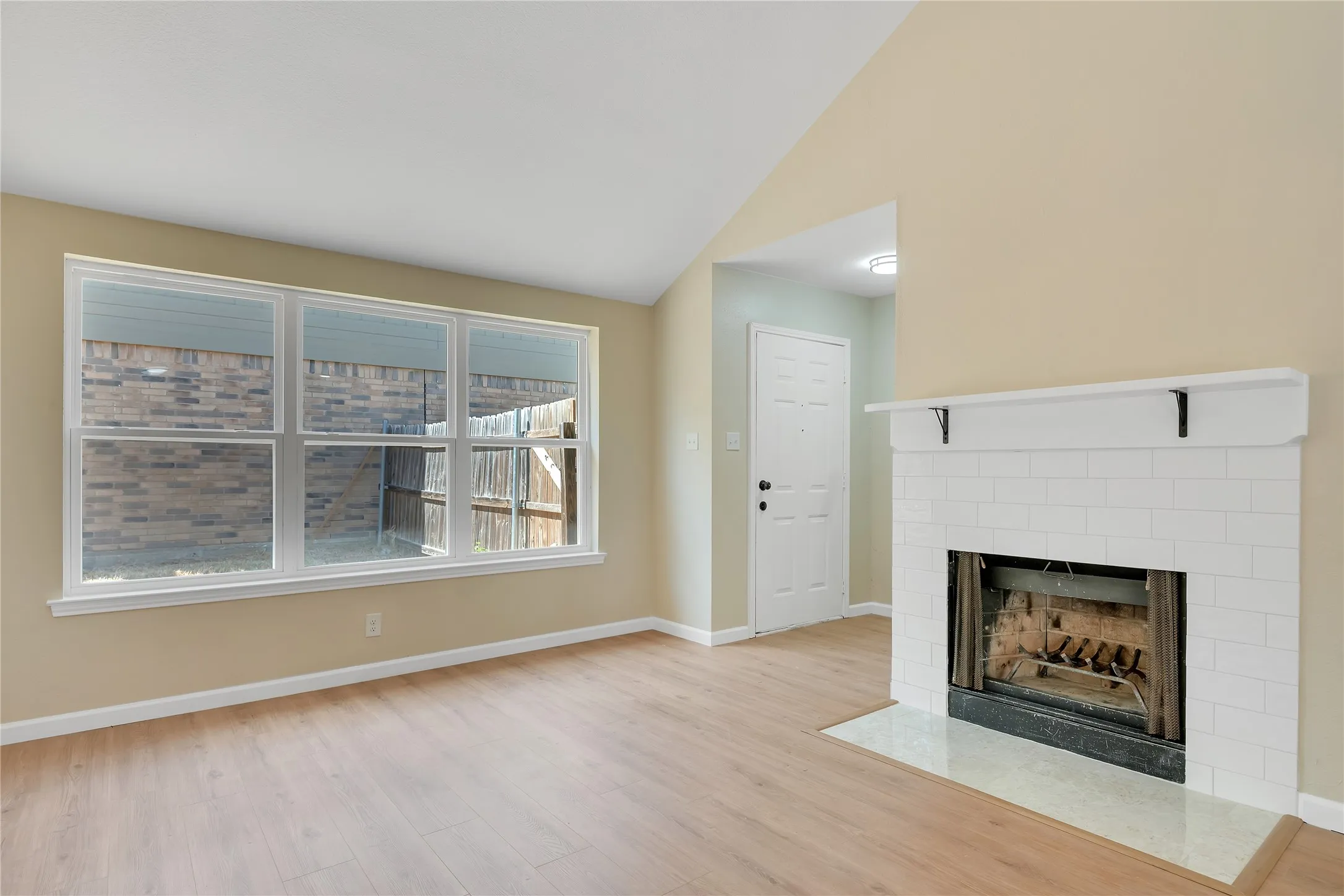 Vaulted ceiling, wood-finish flooring, large multi-pane window, white brick fireplace with mantel, and a white paneled door