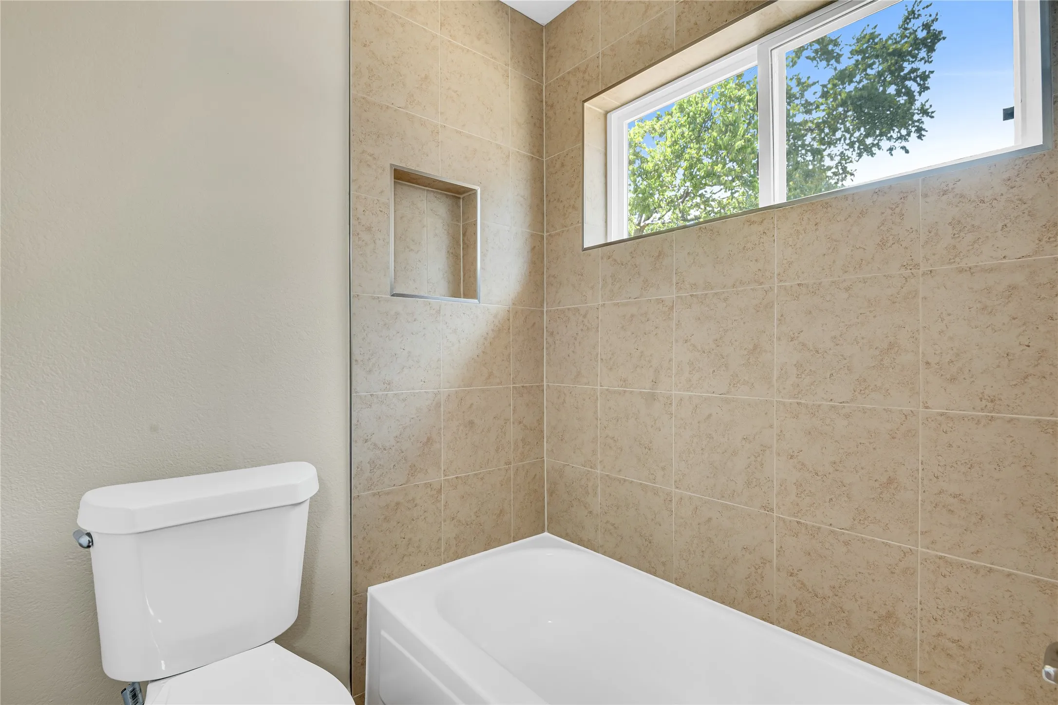 White ceramic bathtub with a tiled surround, recessed shower niche, and a high window