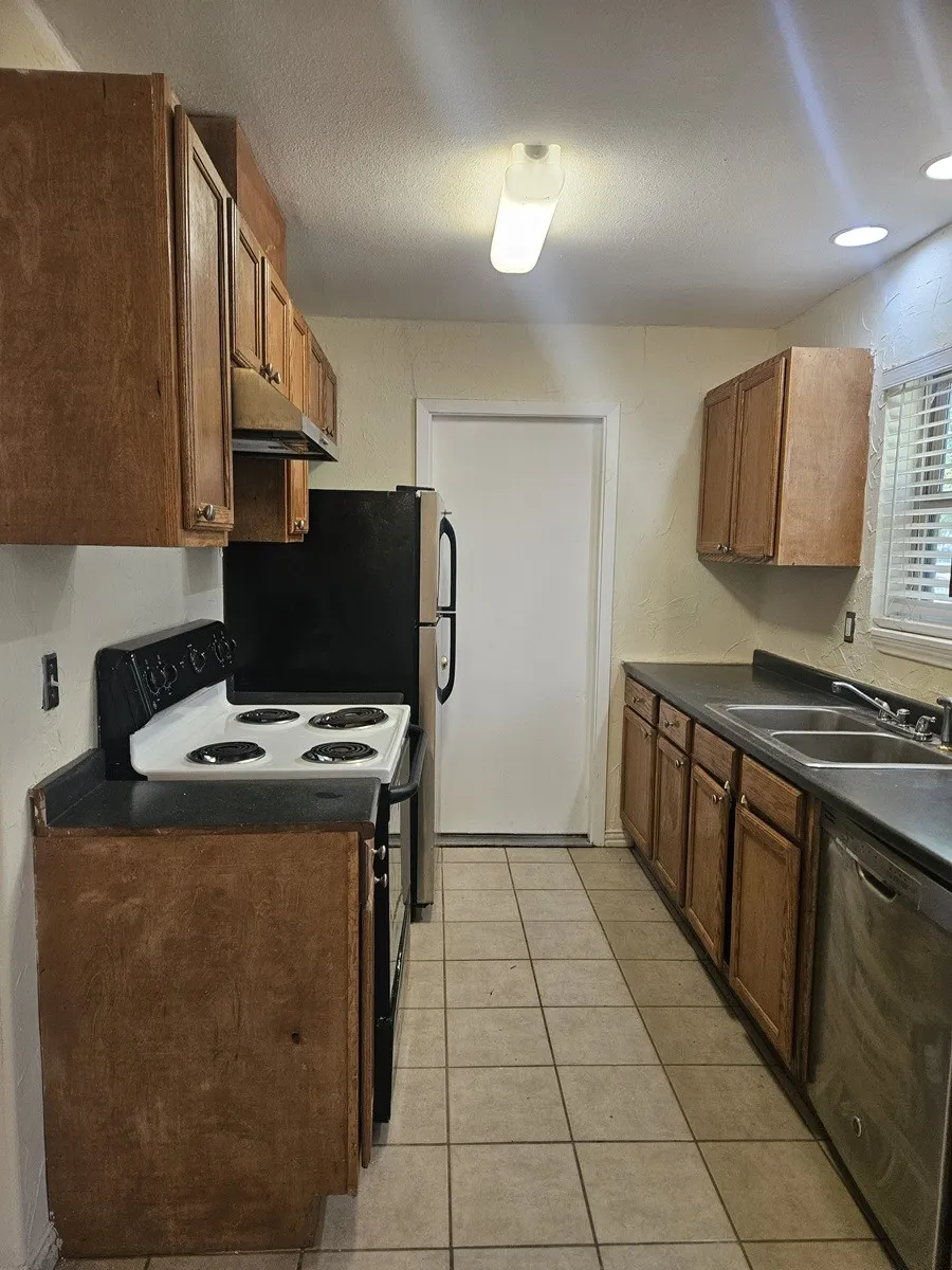 Kitchen featuring wood cabinetry, dark countertops, a stainless steel dishwasher, and tile flooring