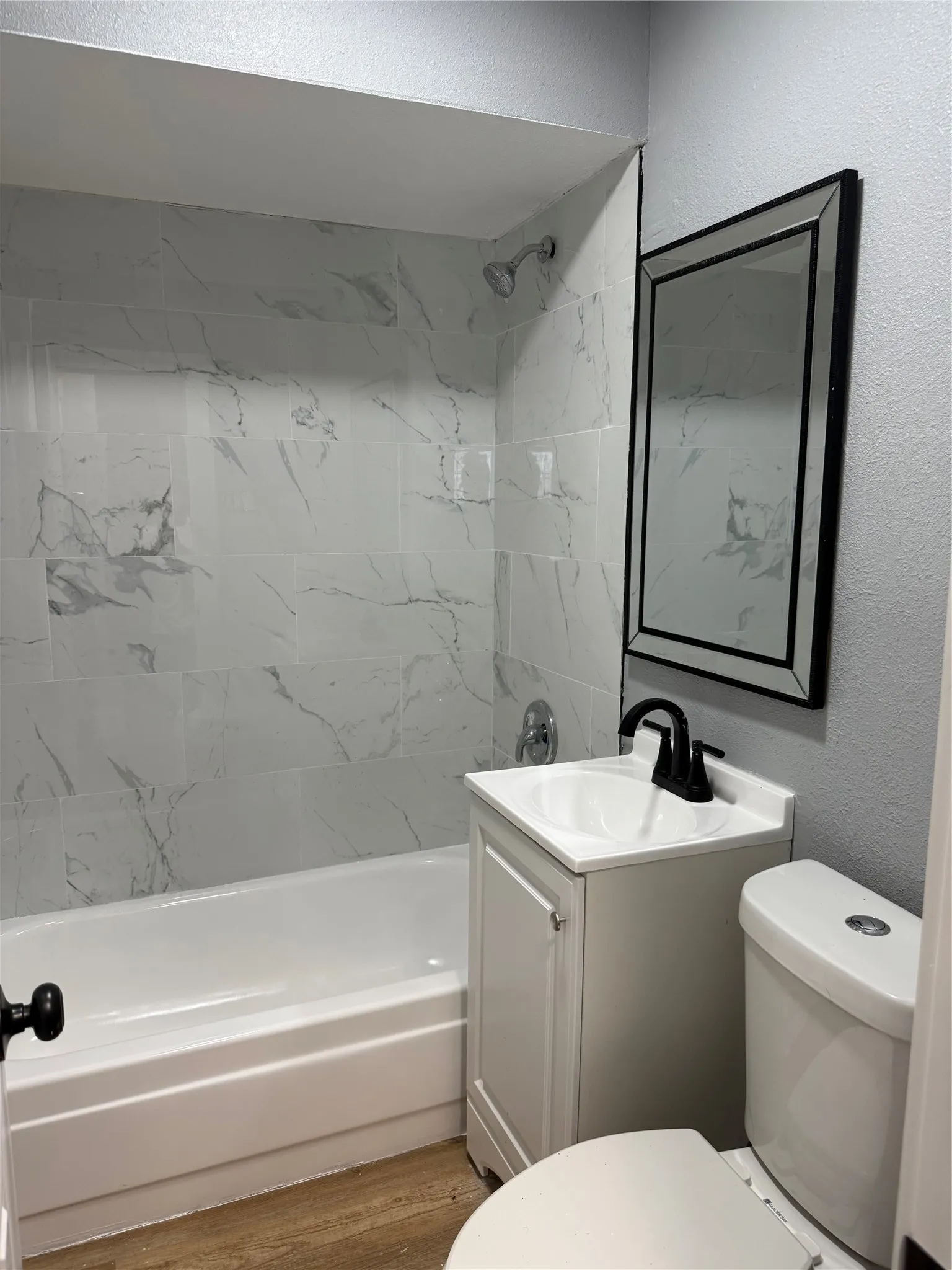 Bathroom featuring a white bathtub with marble-look tile surround, a single vanity with storage, matte black faucet, and wood-finish flooring