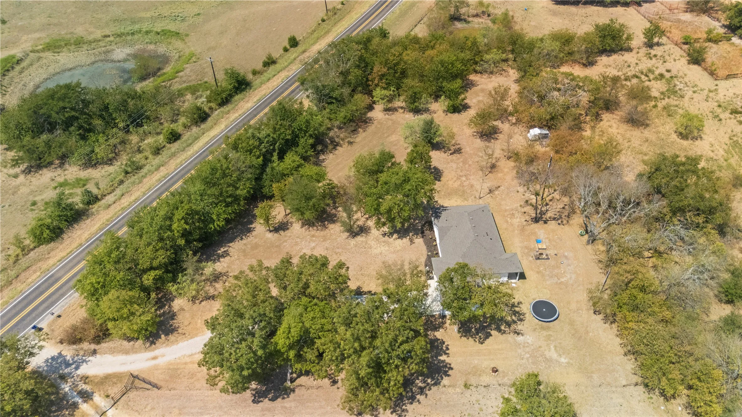 Expansive rural property featuring a single-story residence with a gray roof, mature trees, an unpaved road access, and a visible pond