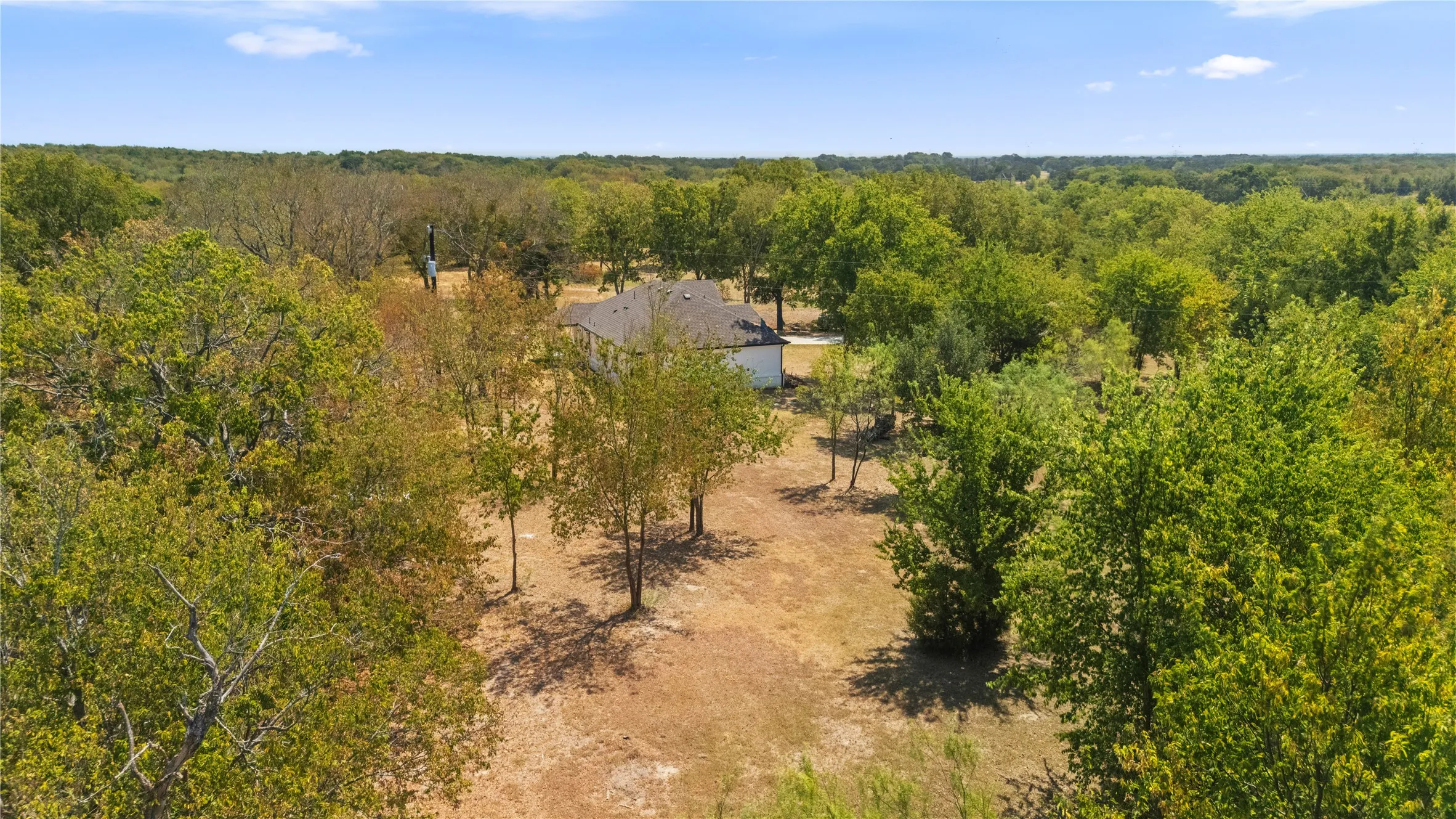 Spacious property featuring a single-story residence with a dark-shingled roof, surrounded by mature trees and natural landscaping