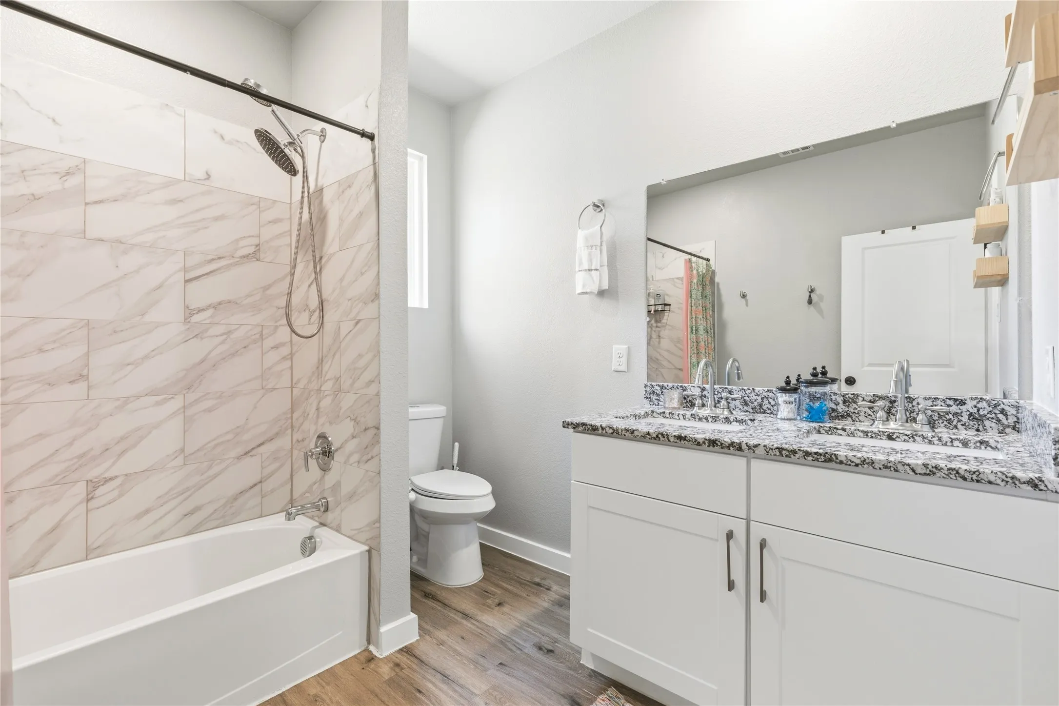 Dual sink vanity with speckled countertop, white cabinetry, wood-finish flooring, white bathtub with marble-style tile surround, and a fixed window
