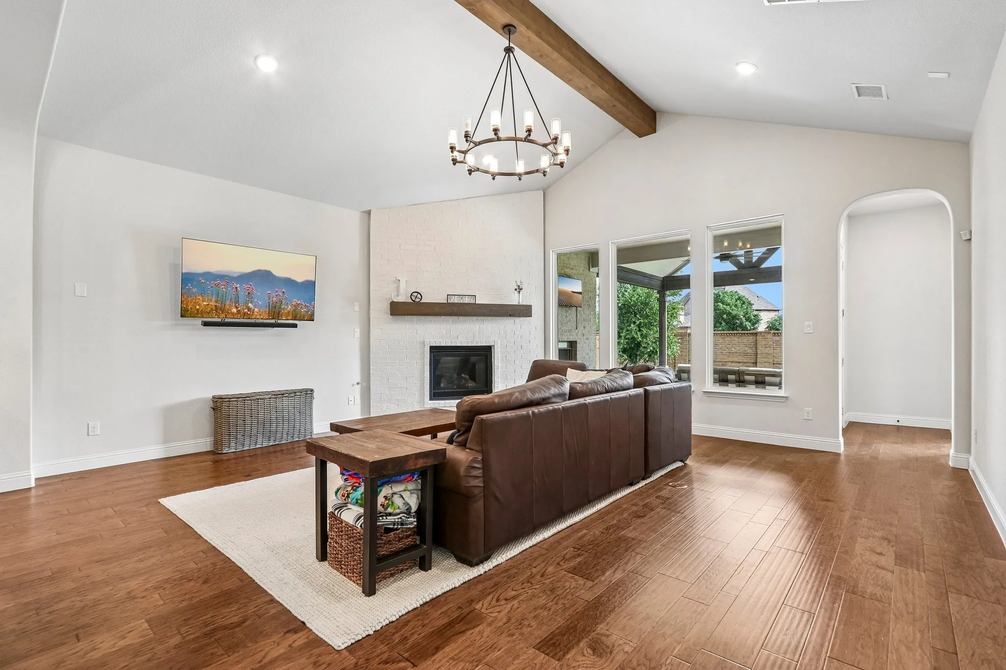 Vaulted ceiling with exposed wood beam, wood-finish flooring, white brick fireplace, arched doorway, and large windows