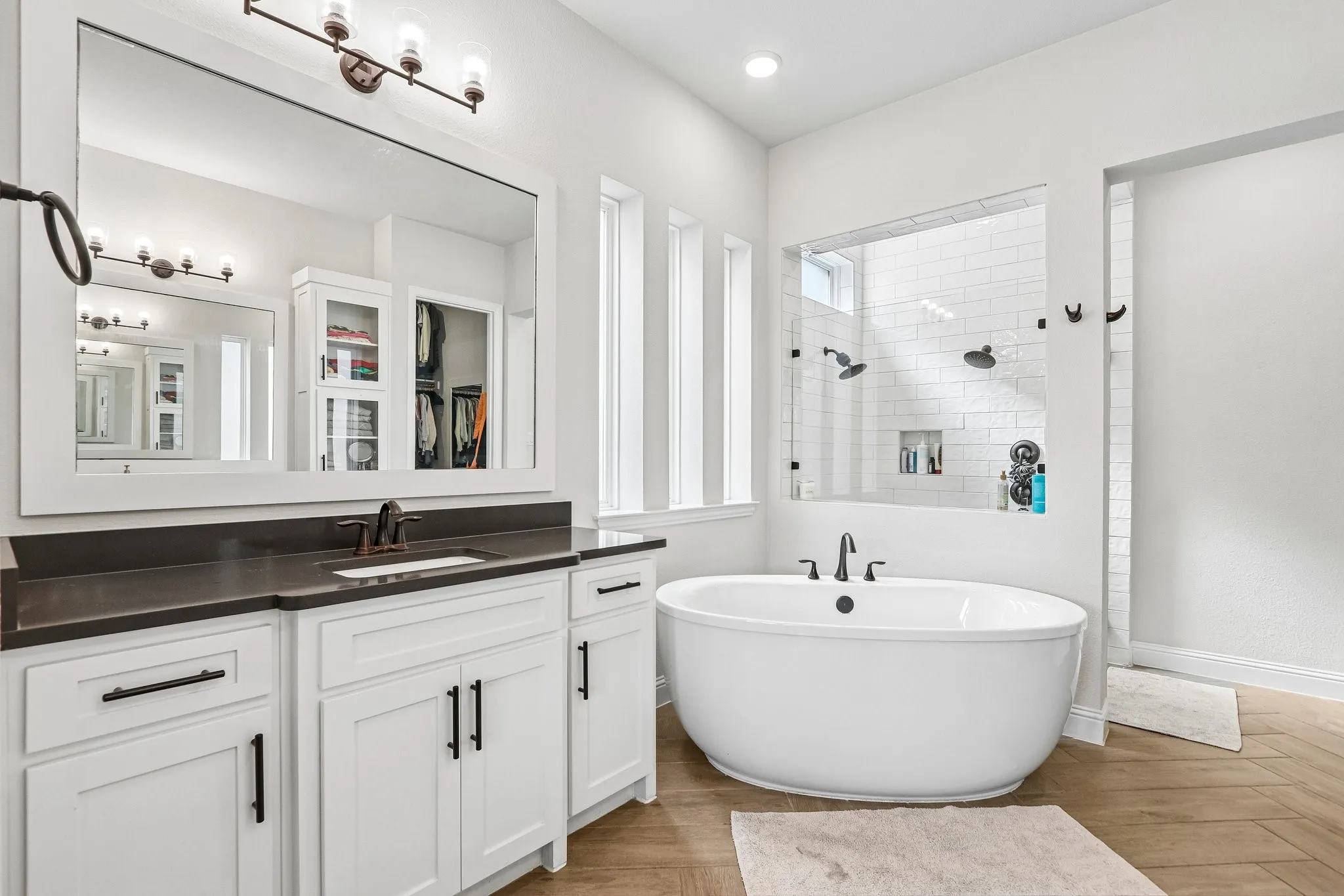 White shaker-style vanity with dark countertop, large framed mirror, freestanding soaking tub, and a glass-enclosed shower with subway tile