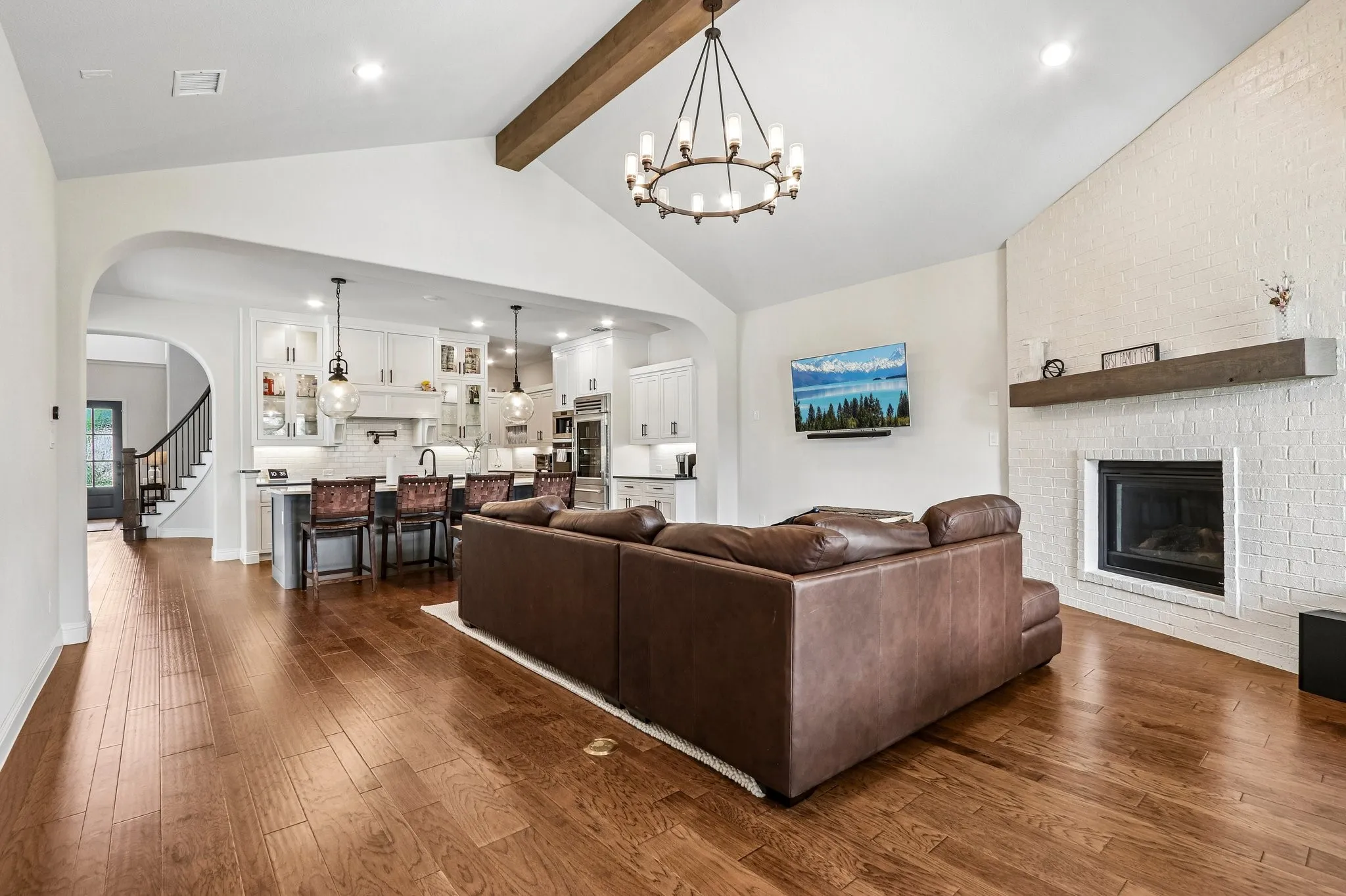 Vaulted ceiling with wood beam, wood-finish flooring, white brick fireplace with mantel, white kitchen cabinetry, and a kitchen island with pendant lighting