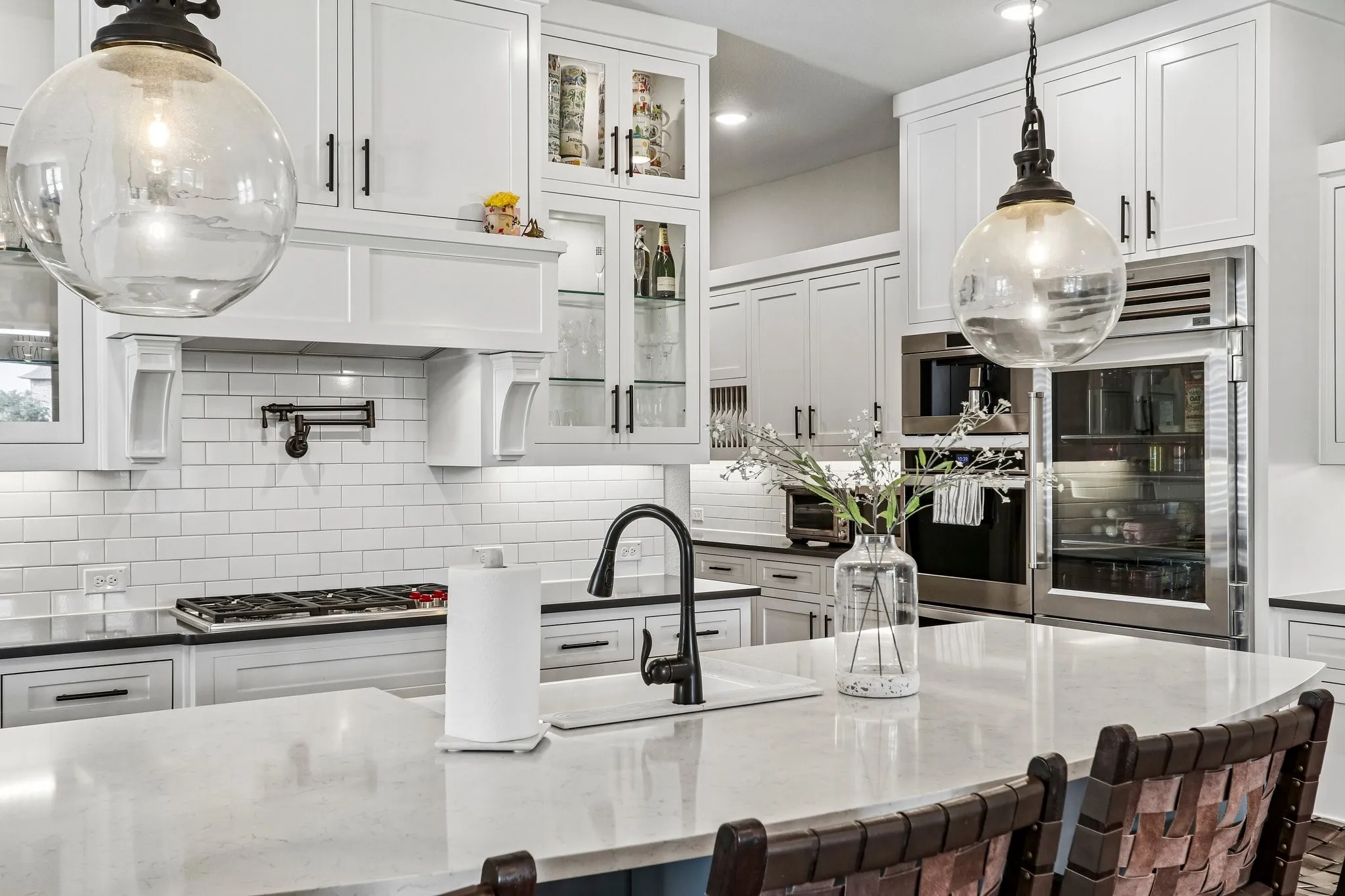 White shaker-style cabinetry with glass-front accents, subway tile backsplash, built-in stainless steel appliances, and a kitchen island with an undermount sink