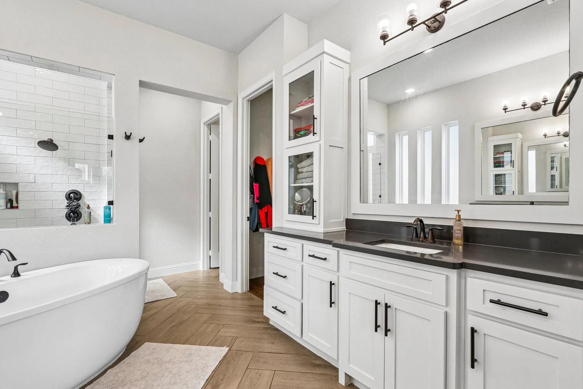 Freestanding soaking tub, walk-in shower with subway tile, dual vanity with undermount sink, dark countertop, and herringbone wood-finish flooring