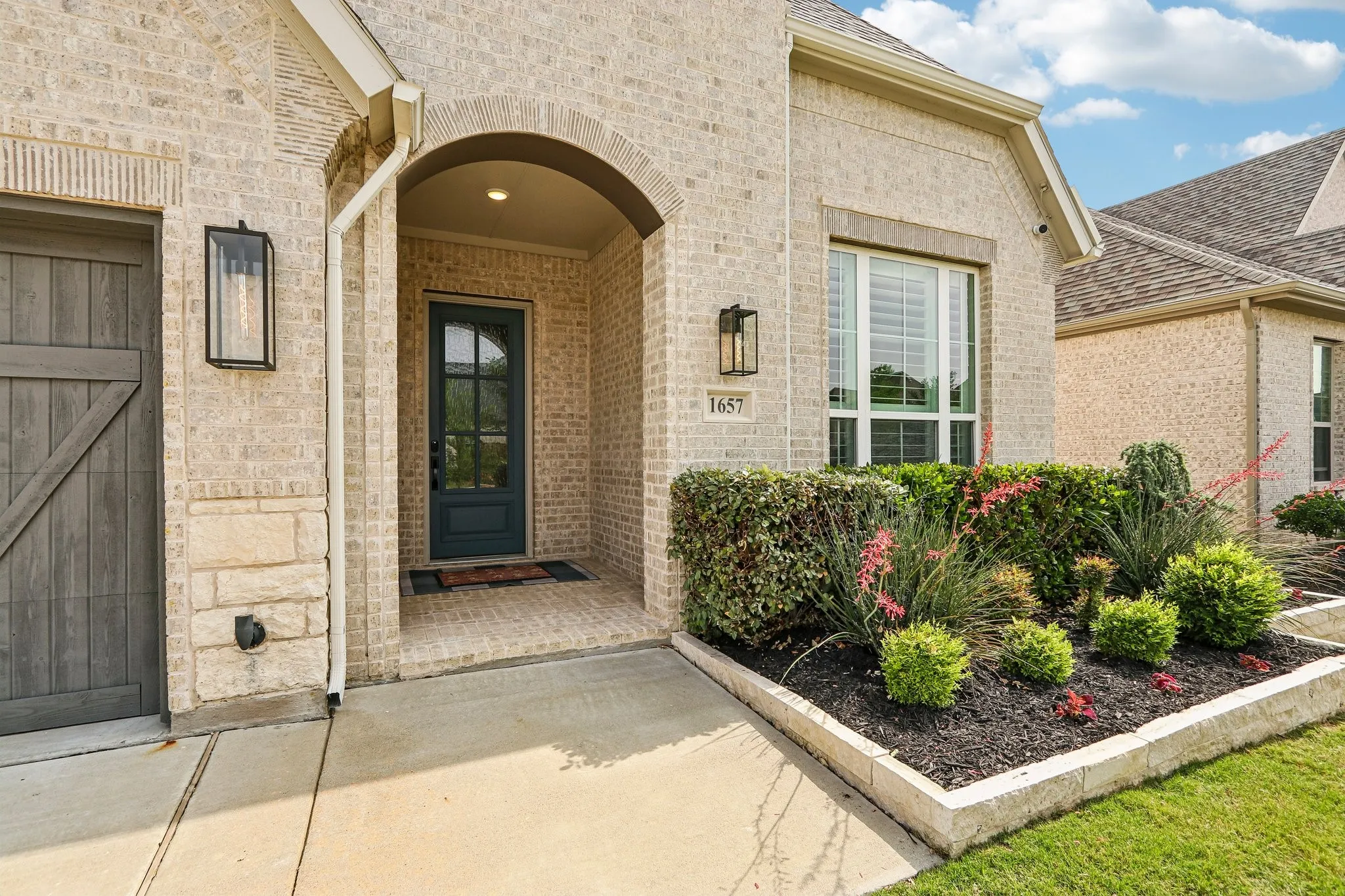 Arched entryway with a dark paneled door, light brick facade, stone accents, and exterior lighting fixtures