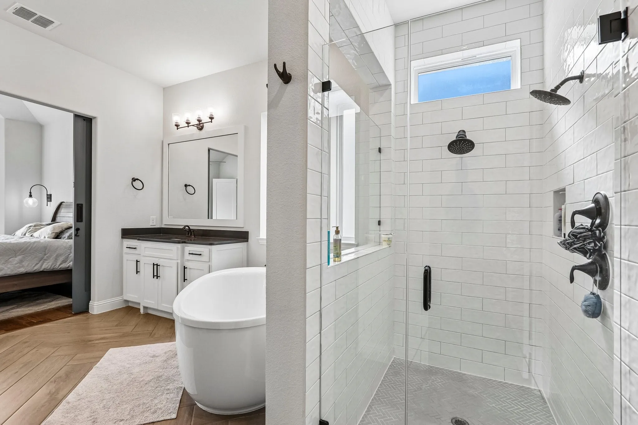 Spacious bathroom featuring a freestanding soaking tub, dual-head shower with subway tile surround, white vanity with dark countertop, and wood-finish flooring