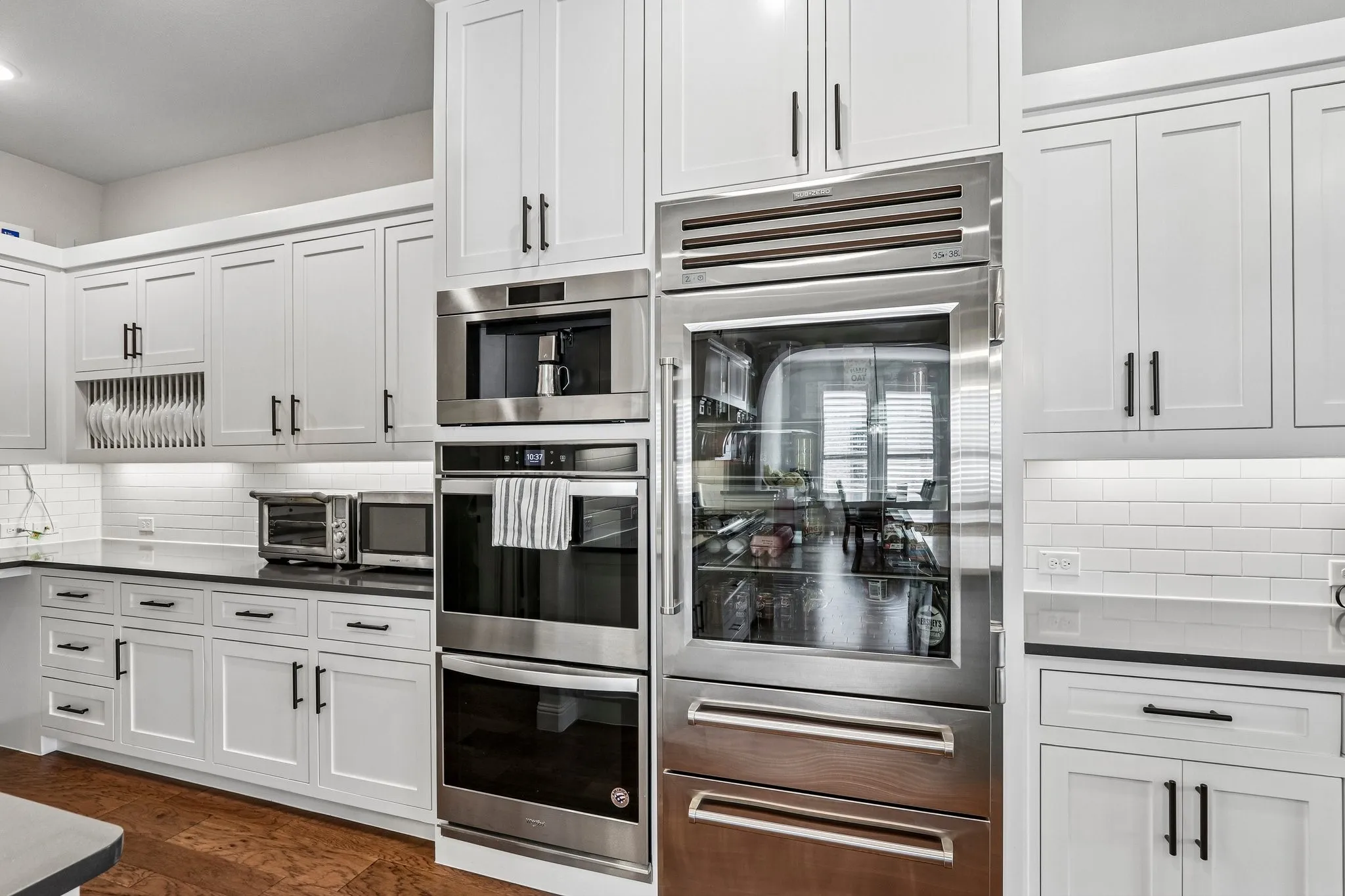 White shaker-style cabinetry with matte black hardware, integrated stainless steel appliances, dark countertops, white subway tile backsplash, and wood-finish flooring