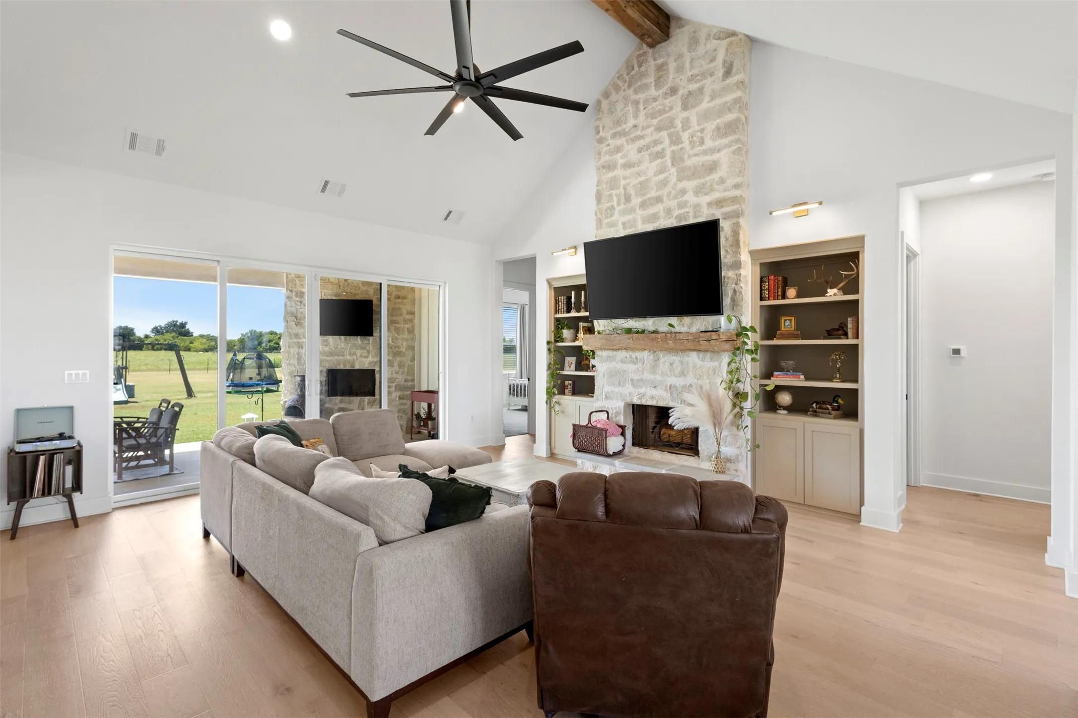 Vaulted ceiling with exposed wood beam, stone fireplace with wood mantle, built-in shelving, wood-finish flooring, and sliding glass doors to outdoor patio