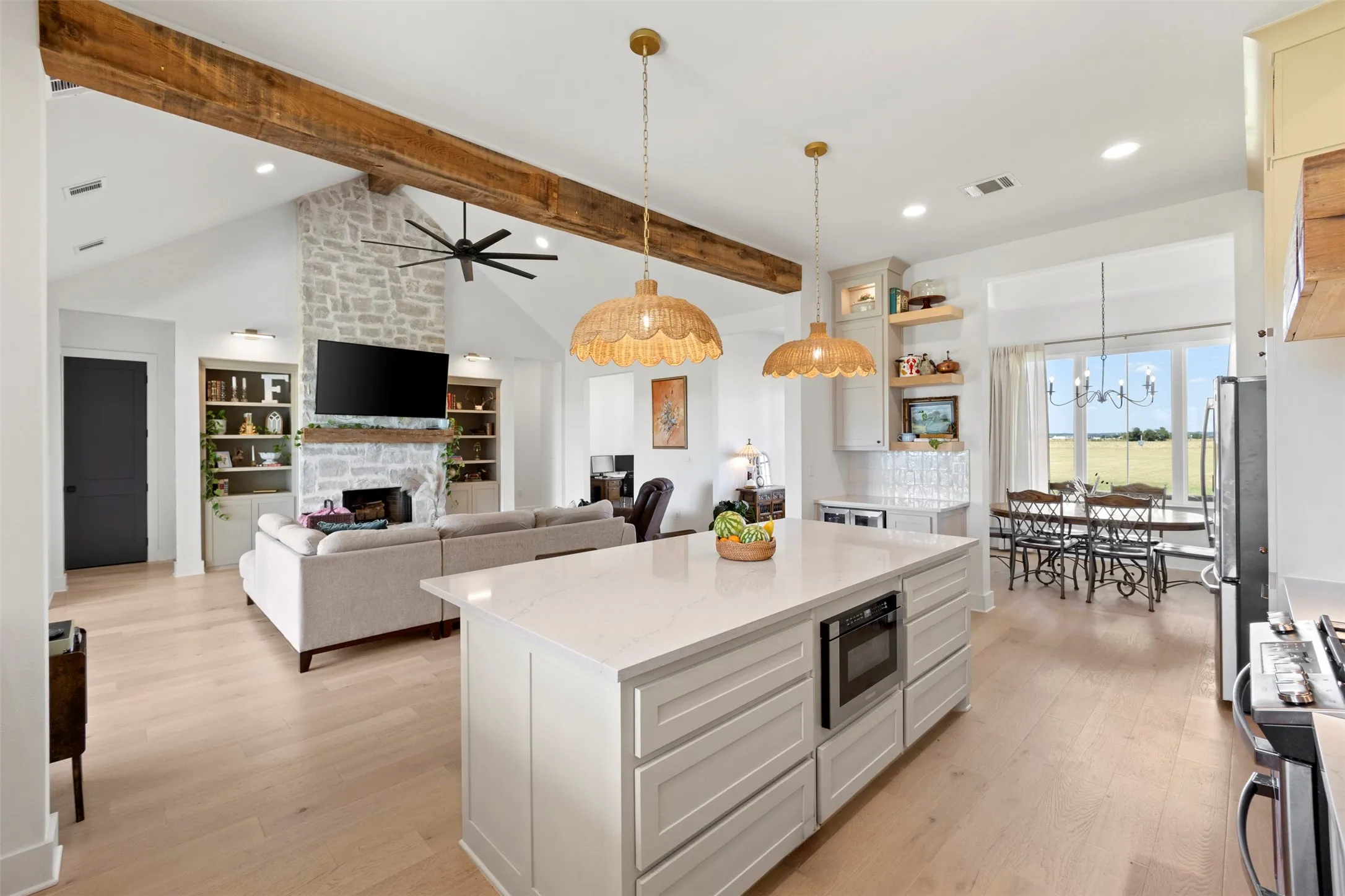 Open-concept living space with vaulted ceilings, exposed wood beams, a stone fireplace, wood-finish flooring, and a kitchen island with built-in microwave