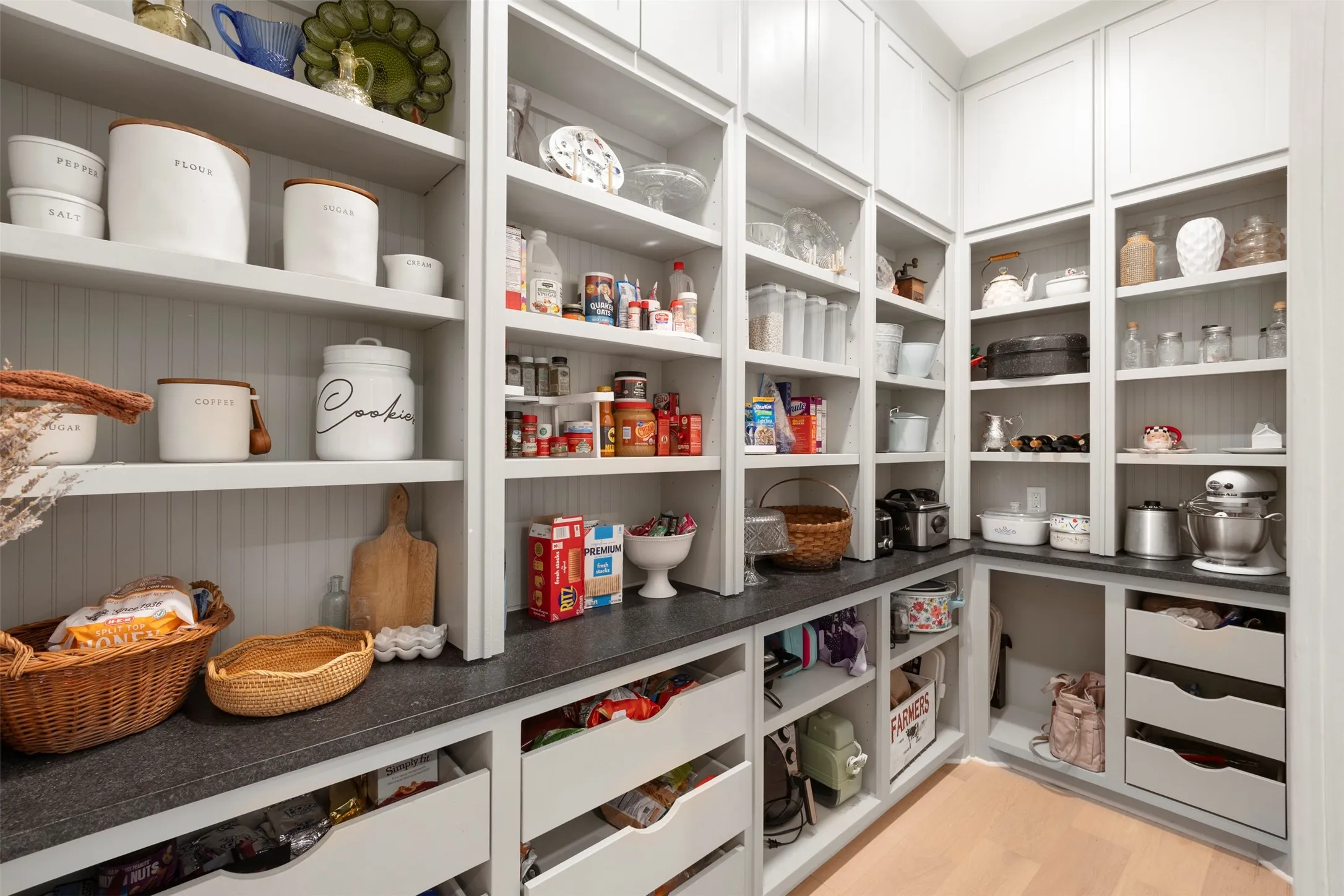 Spacious walk-in pantry featuring extensive open shelving, dark countertops, multiple storage drawers, white cabinetry, and light wood-finish flooring