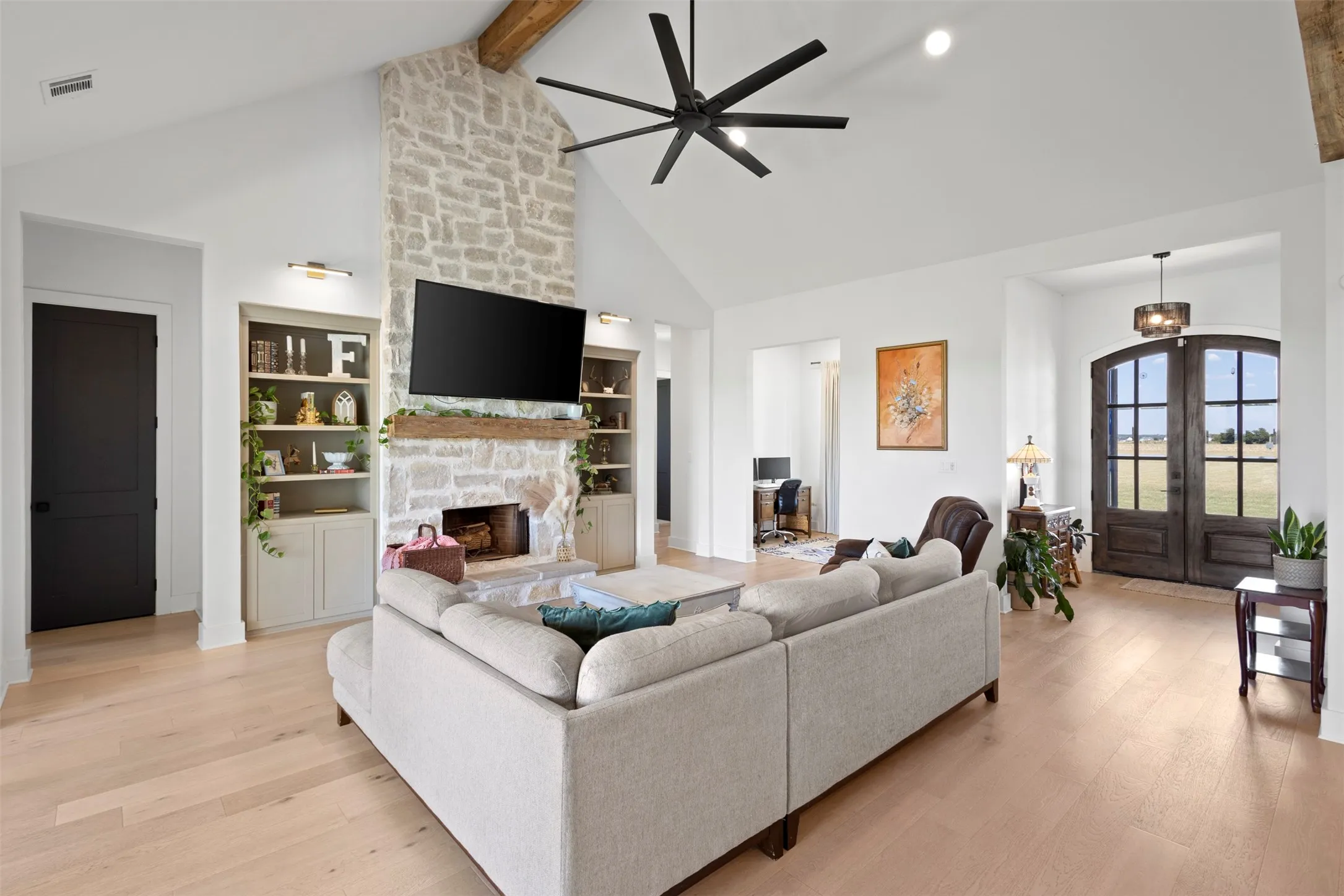 Vaulted ceiling with exposed wood beam, stone fireplace with wood mantel, built-in shelving, wood-finish flooring, and arched double entry doors