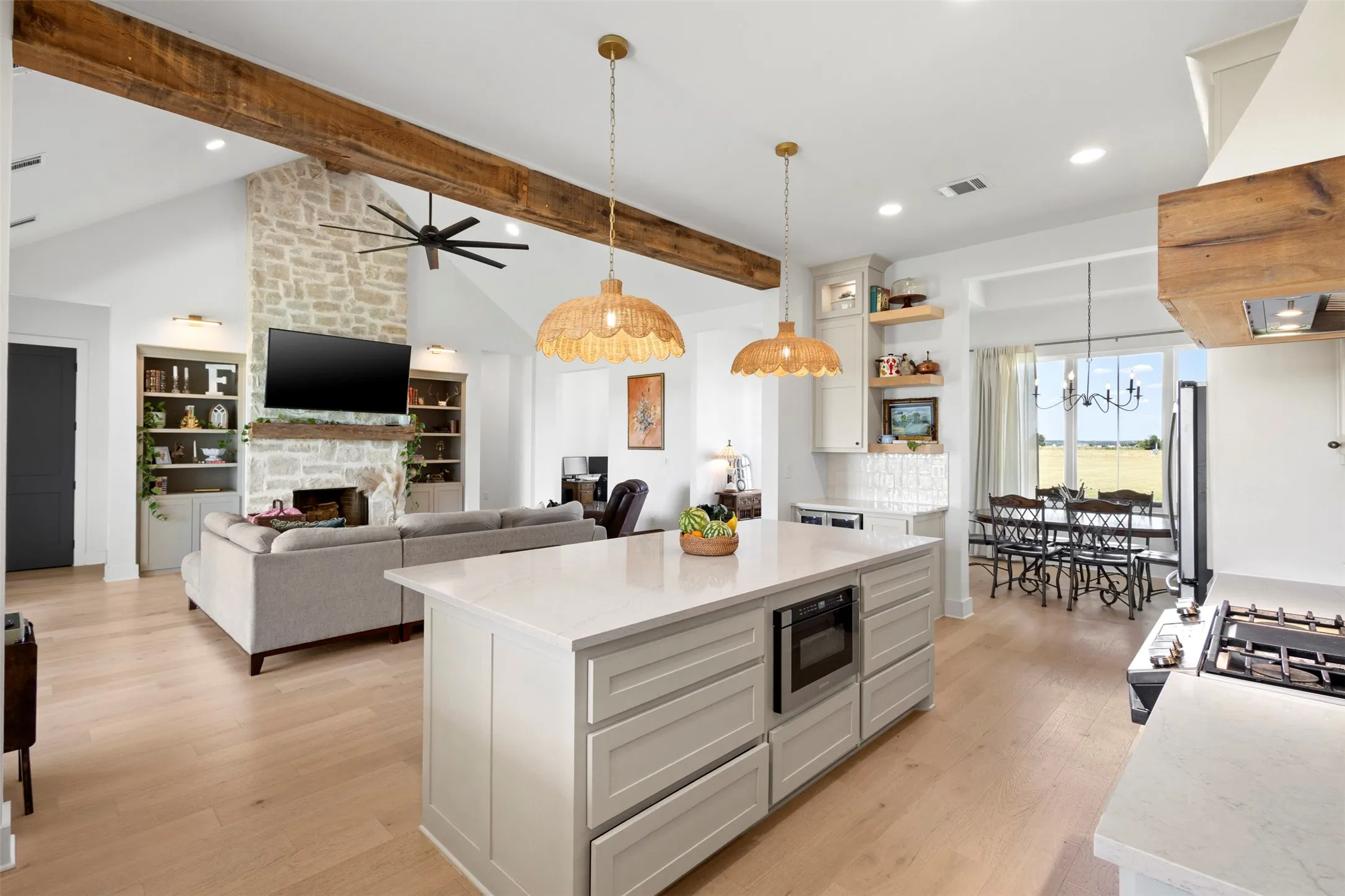 Vaulted ceiling with exposed wood beams, stone-clad fireplace with mantel, built-in shelving, center island with integrated microwave, and wood-finish flooring