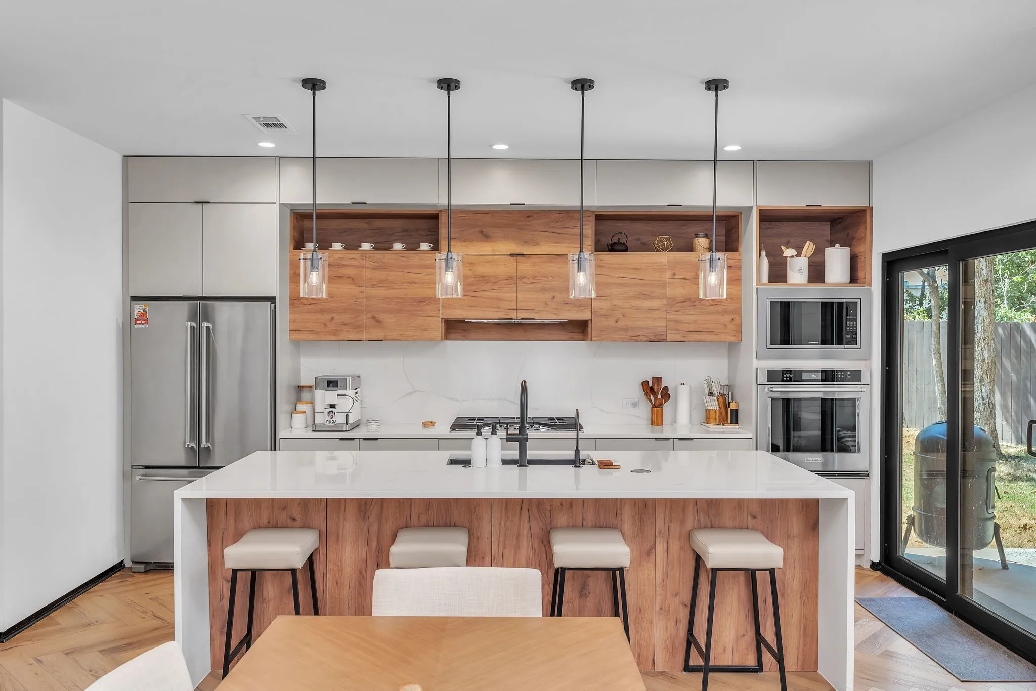 Expansive kitchen island with a waterfall countertop, four pendant lights, stainless steel appliances, wood-finish cabinetry, and herringbone wood-finish flooring