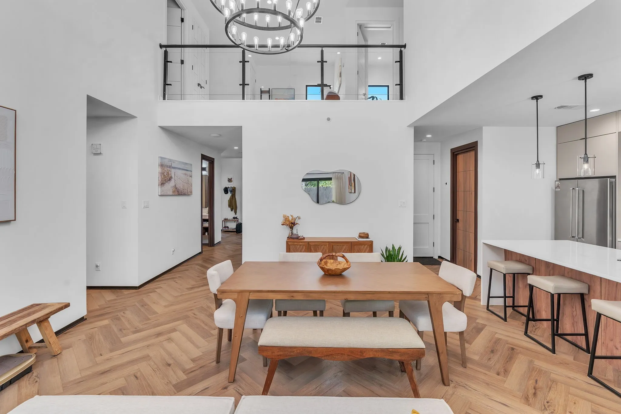 Two-story living space featuring a second-floor walkway with glass panel railing, a contemporary chandelier, herringbone wood-finish flooring, and a kitchen island with a white countertop