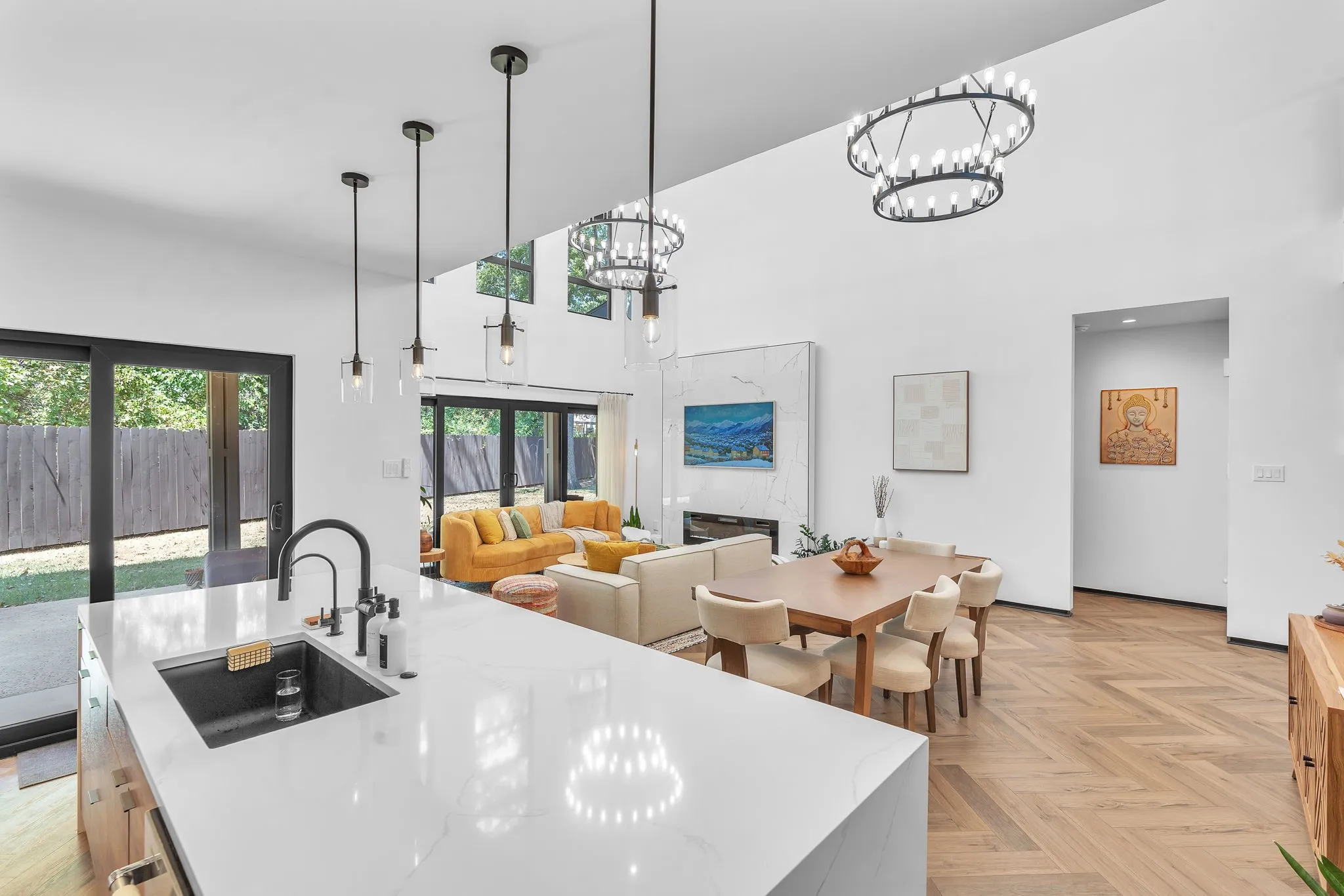 Vaulted ceilings with layered chandeliers, wood-finish herringbone flooring, white countertop kitchen island with integrated sink, and an elegant fireplace with stone surround