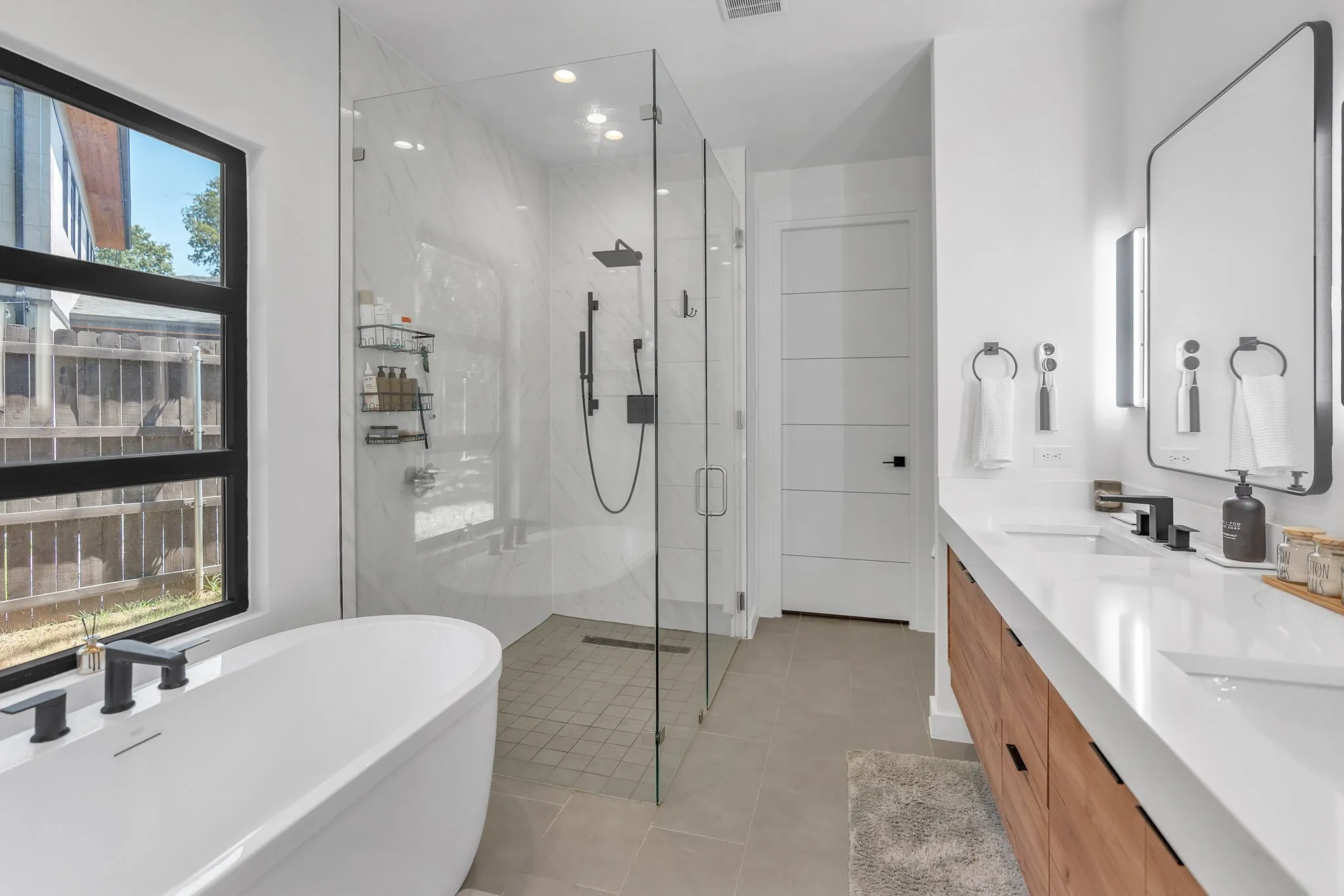 Freestanding soaking tub with matte black fixtures, glass-enclosed shower with tile surround, dual vanity with undermount sinks and wood-finish cabinetry