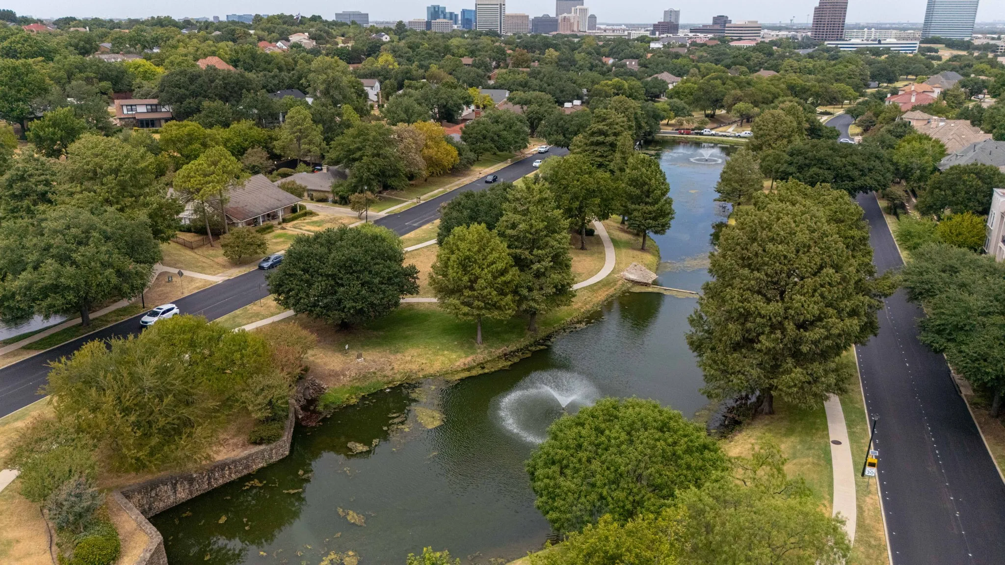 Expansive aerial perspective showcasing a neighborhood with mature trees, a tranquil pond with a fountain, and paved pathways