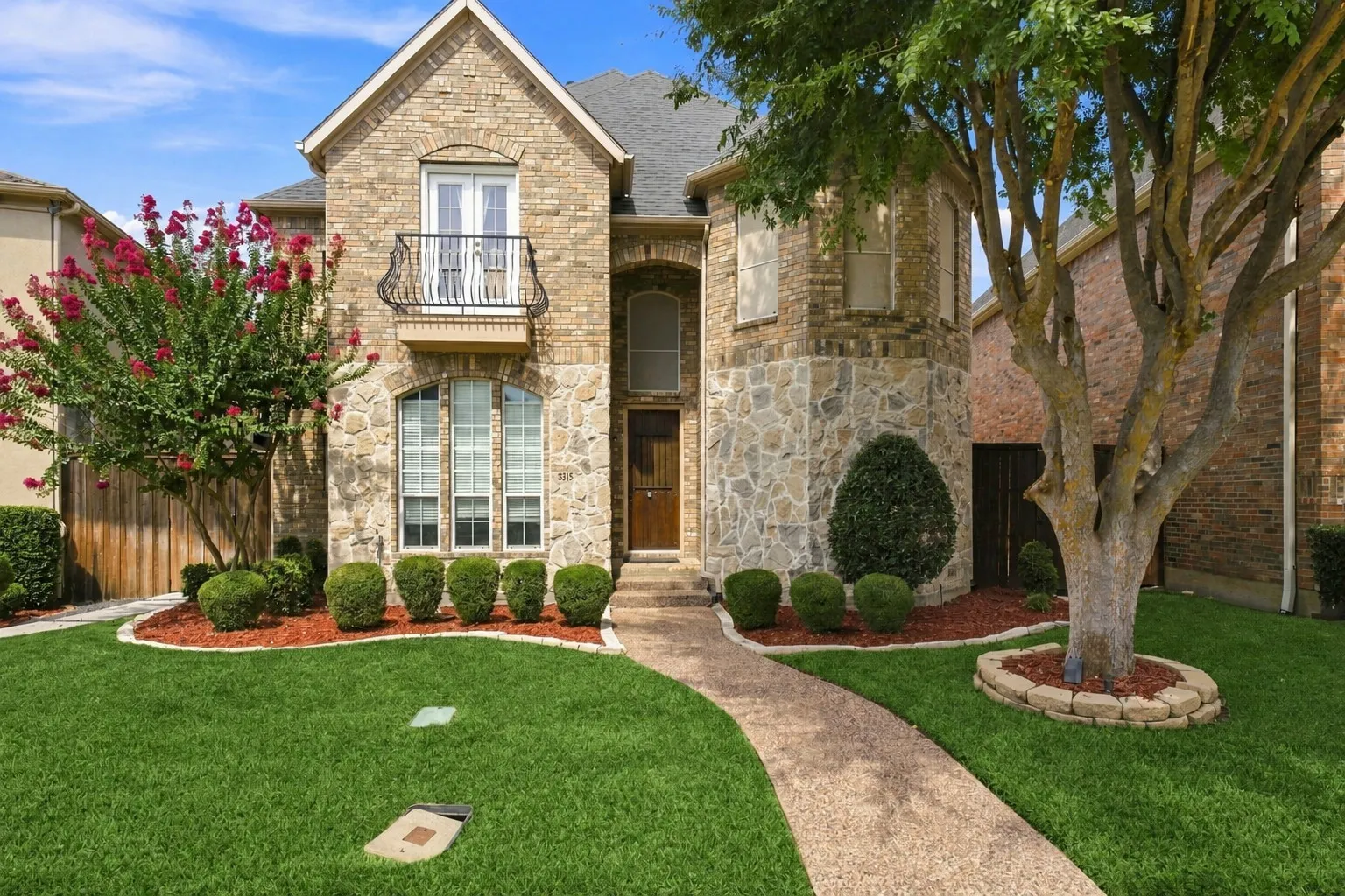 Brick and stone facade with a Juliet balcony, mature landscaping, and a curved entry walkway