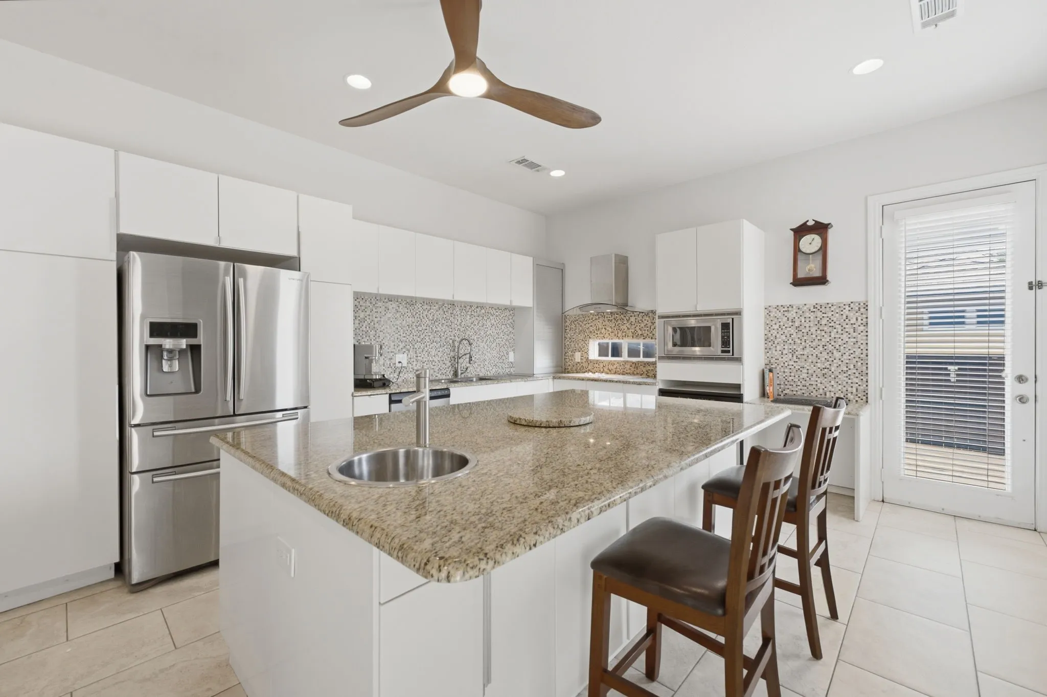 Spacious kitchen featuring an expansive granite-top island with an integrated sink, stainless steel appliances, white cabinetry, and a mosaic tile backsplash