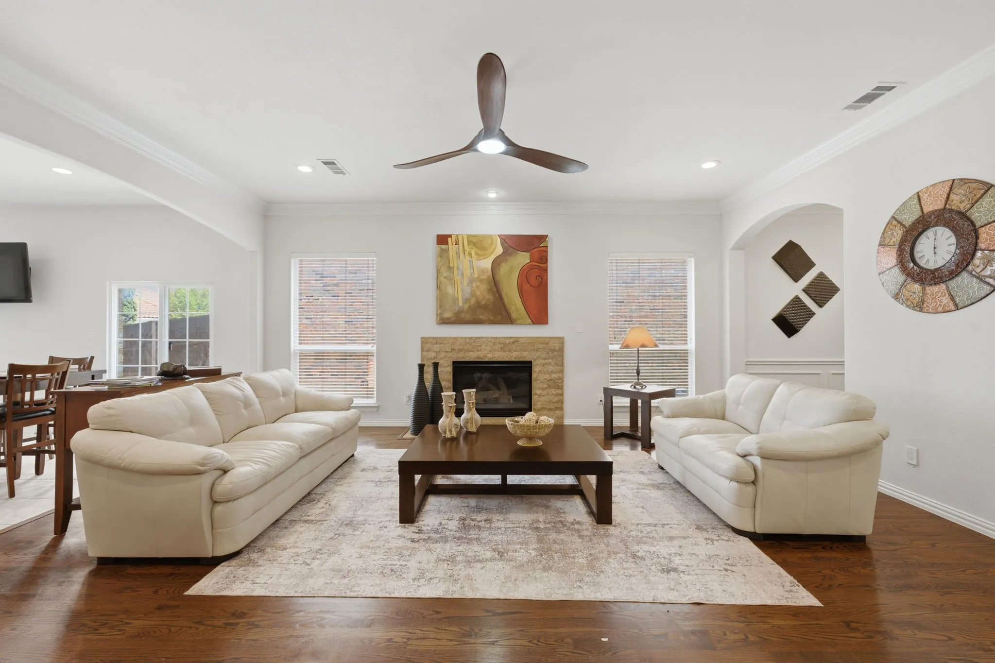 Spacious living area featuring wood-finish flooring, a stone-surround fireplace, crown molding, a ceiling fan, and recessed lighting
