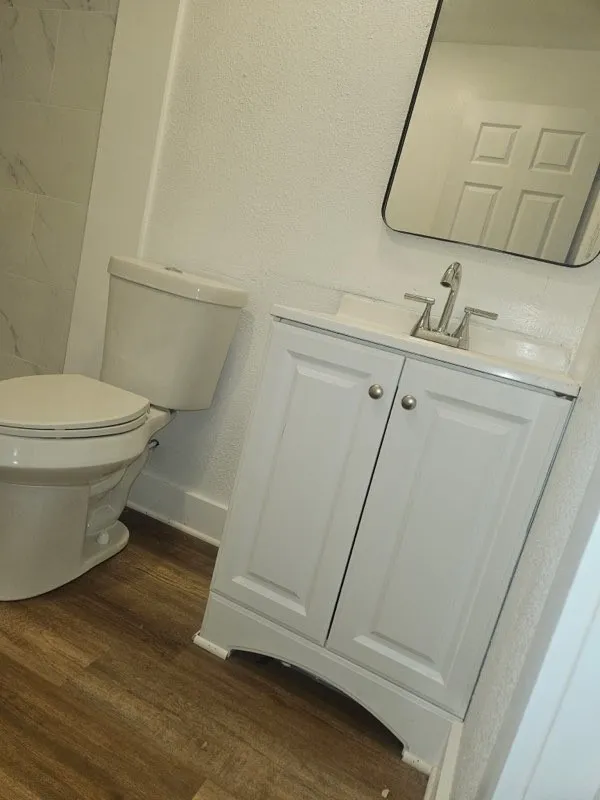 White vanity with dual cabinet storage, wood-finish flooring, a contemporary mirror, and a white toilet