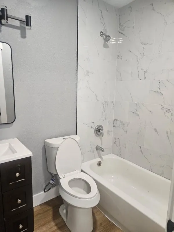 Bathroom featuring a full tub with shower, white marbled tile surround, dark wood-finish vanity with white countertop, and wood-finish flooring