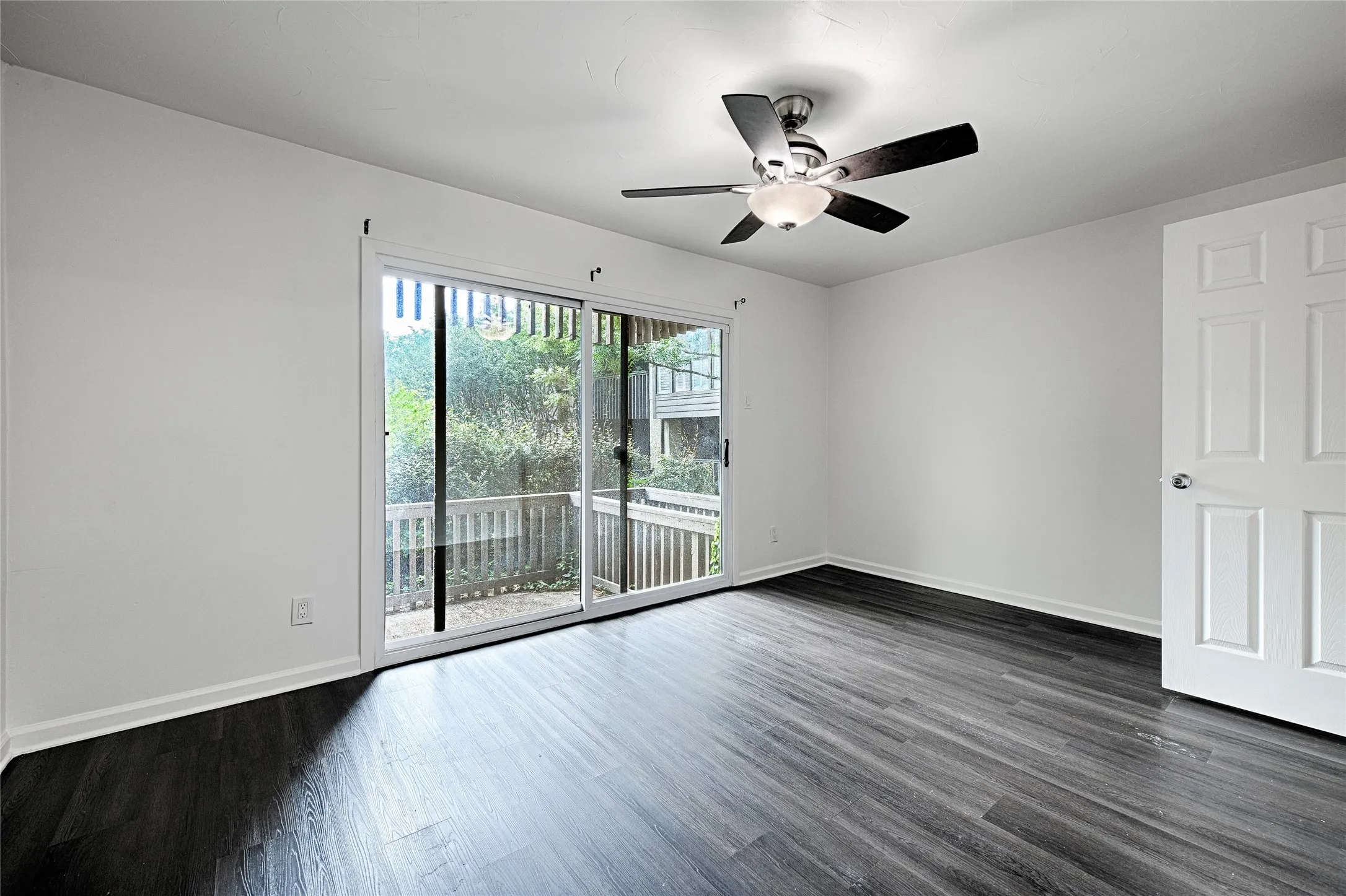 Wood-finish flooring, sliding glass door, private balcony with wood railing, ceiling fan with integrated lighting, and a paneled interior door