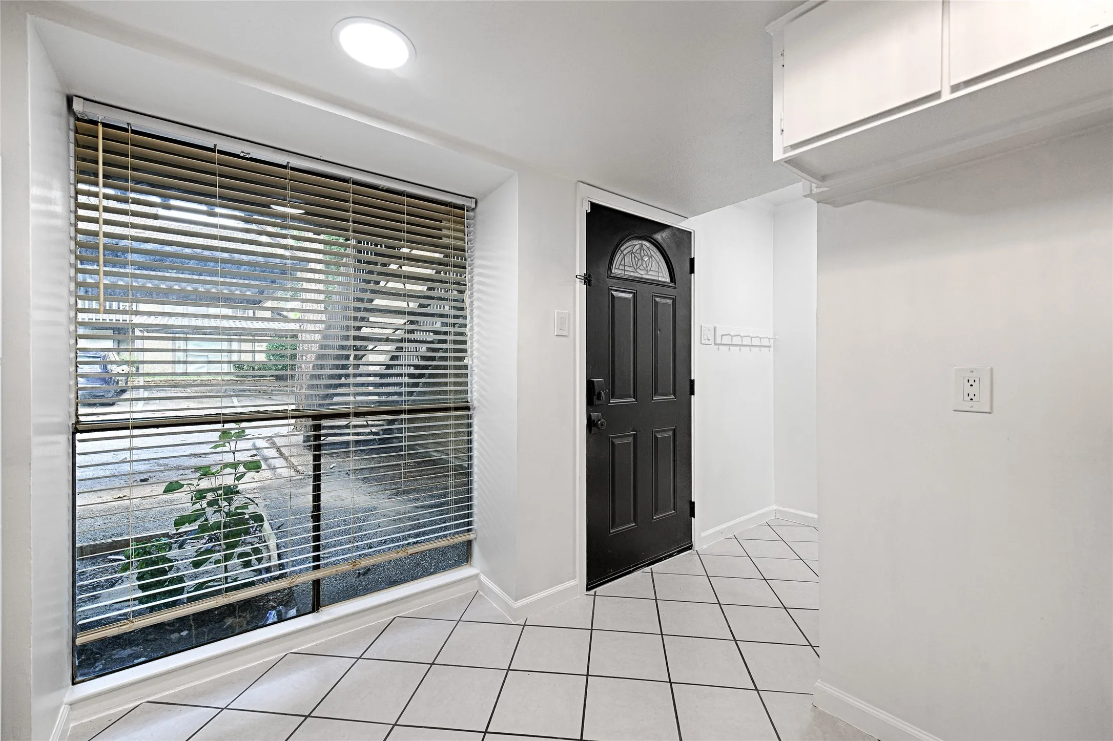 Tiled flooring, recessed ceiling lighting, a dark-paneled entry door with an arched decorative glass insert, a large window with blinds, and white built-in overhead cabinetry