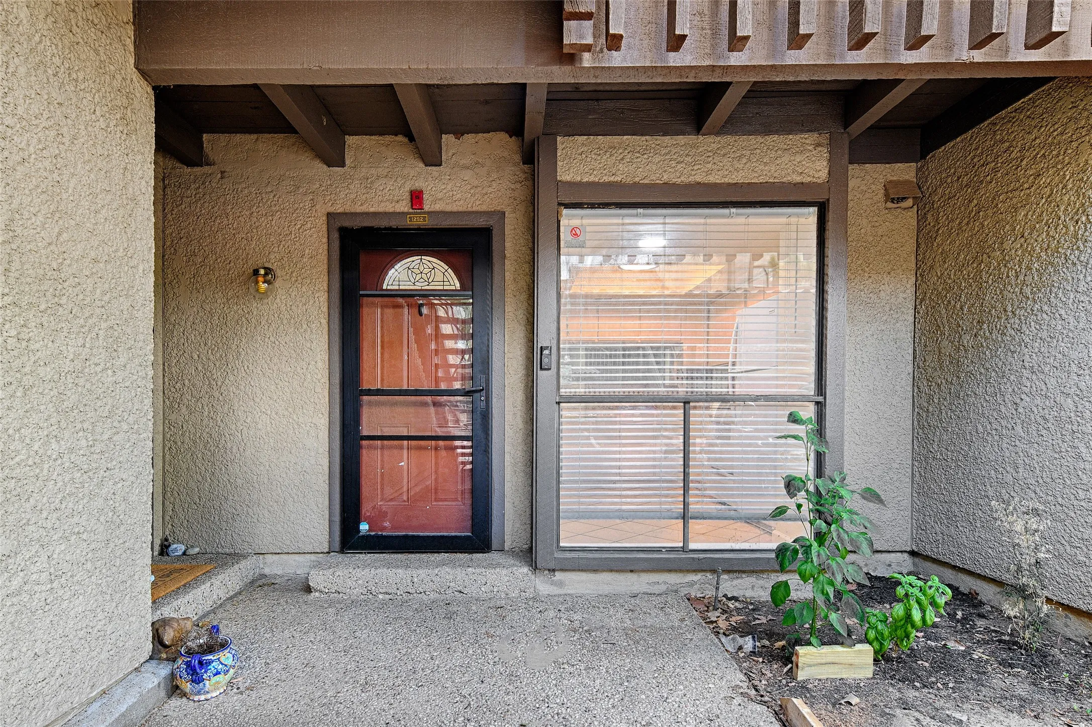 Entryway featuring a red paneled door with decorative glass, a full-length screen door, an adjacent window with horizontal blinds, and a textured stucco exterior