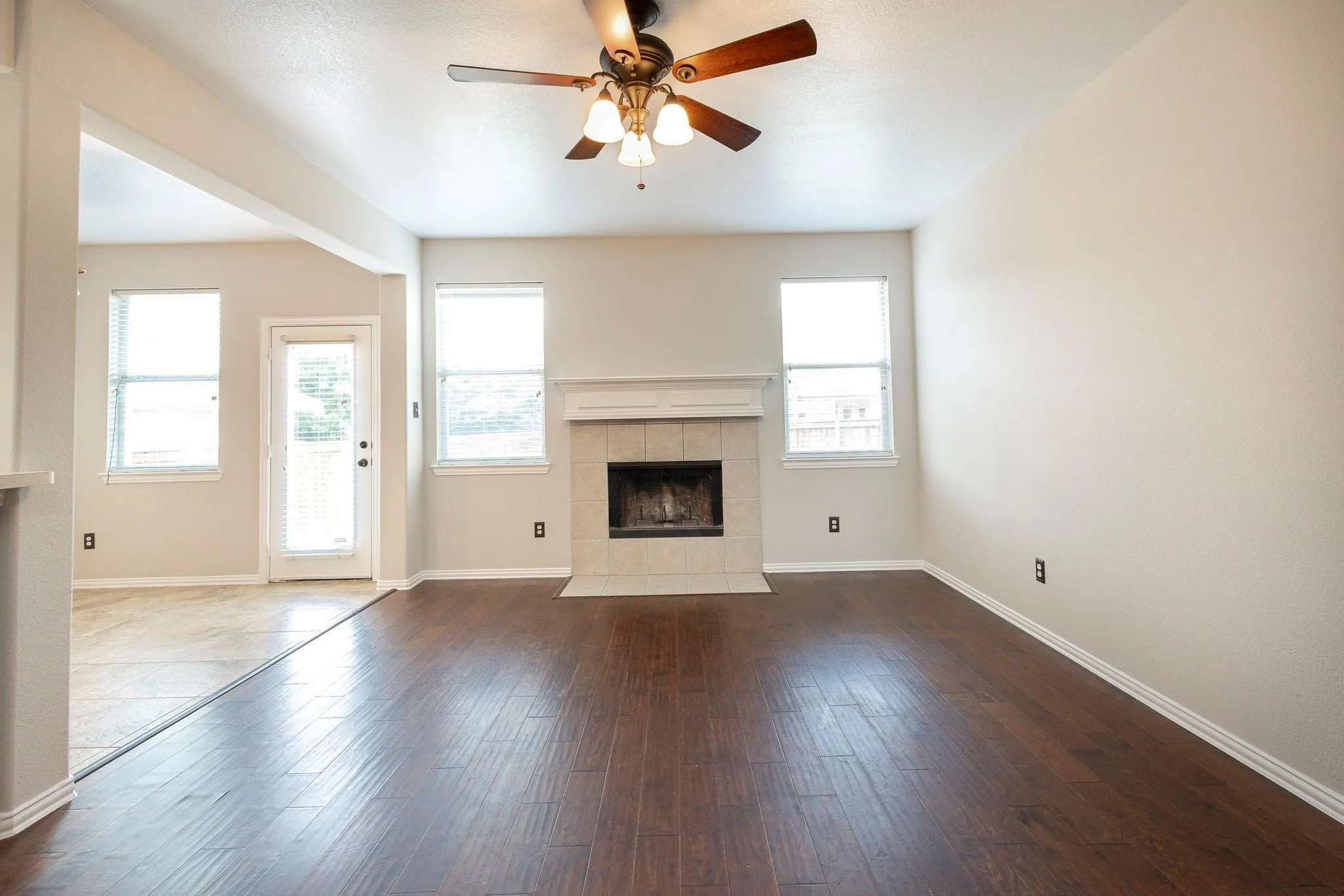 Wood-finish flooring, ceiling fan, fireplace with tile surround and mantel, multiple windows with blinds, and a glass-paneled exterior door