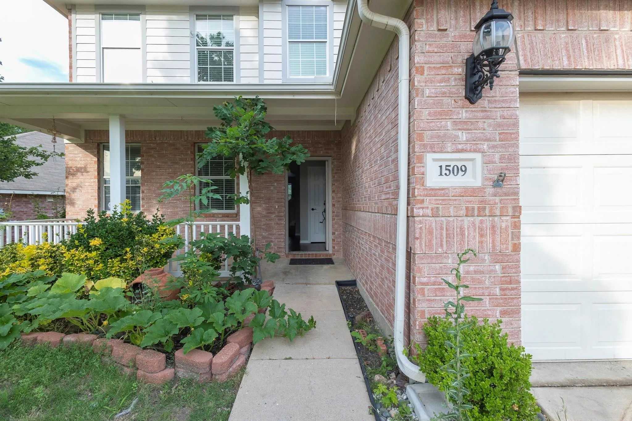 Brick exterior with covered porch, white railing, mature landscaping, concrete walkway, and garage door
