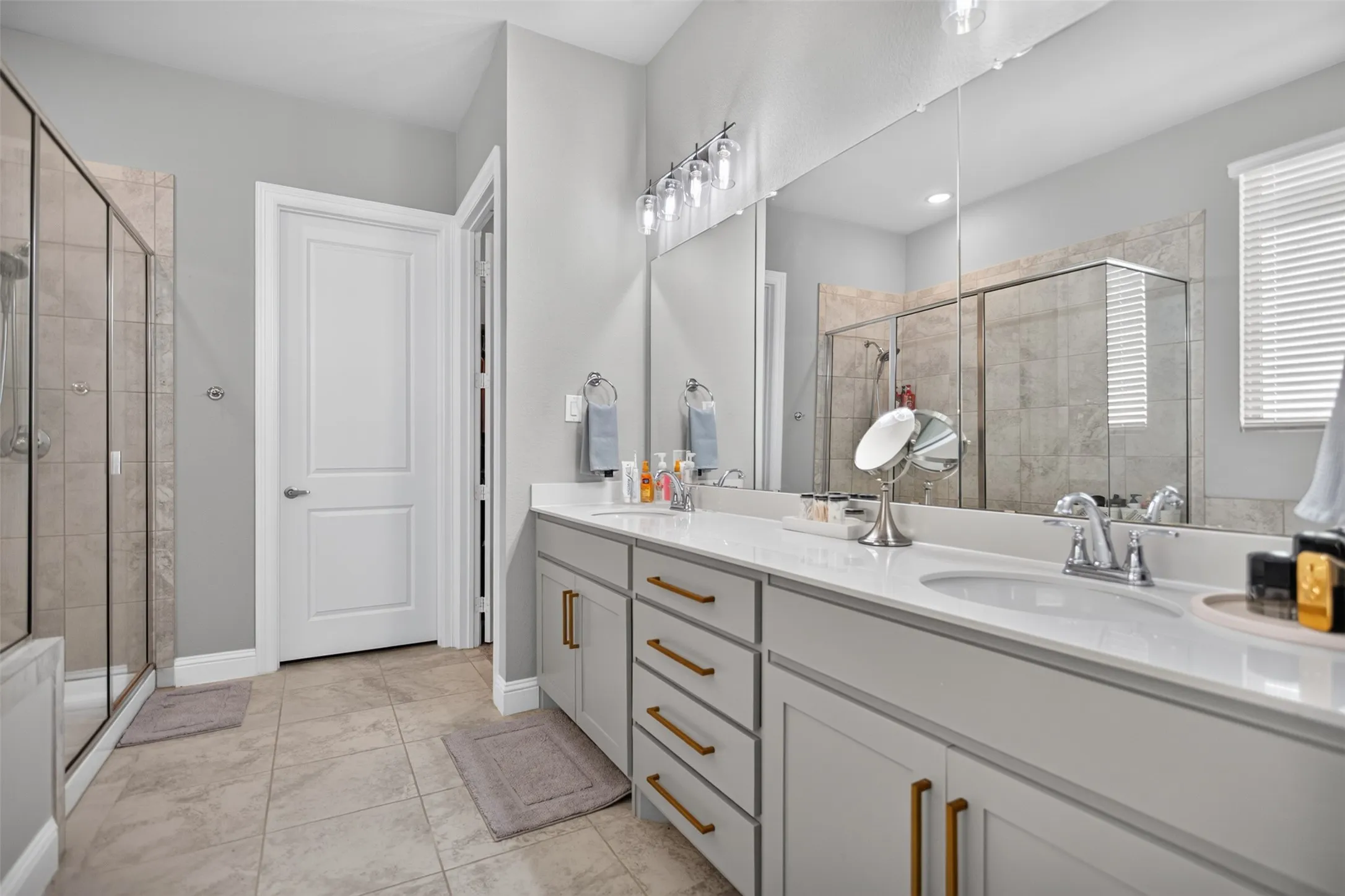 Dual vanity with white countertops, light gray cabinetry, gold-tone hardware, and a framed glass shower enclosure
