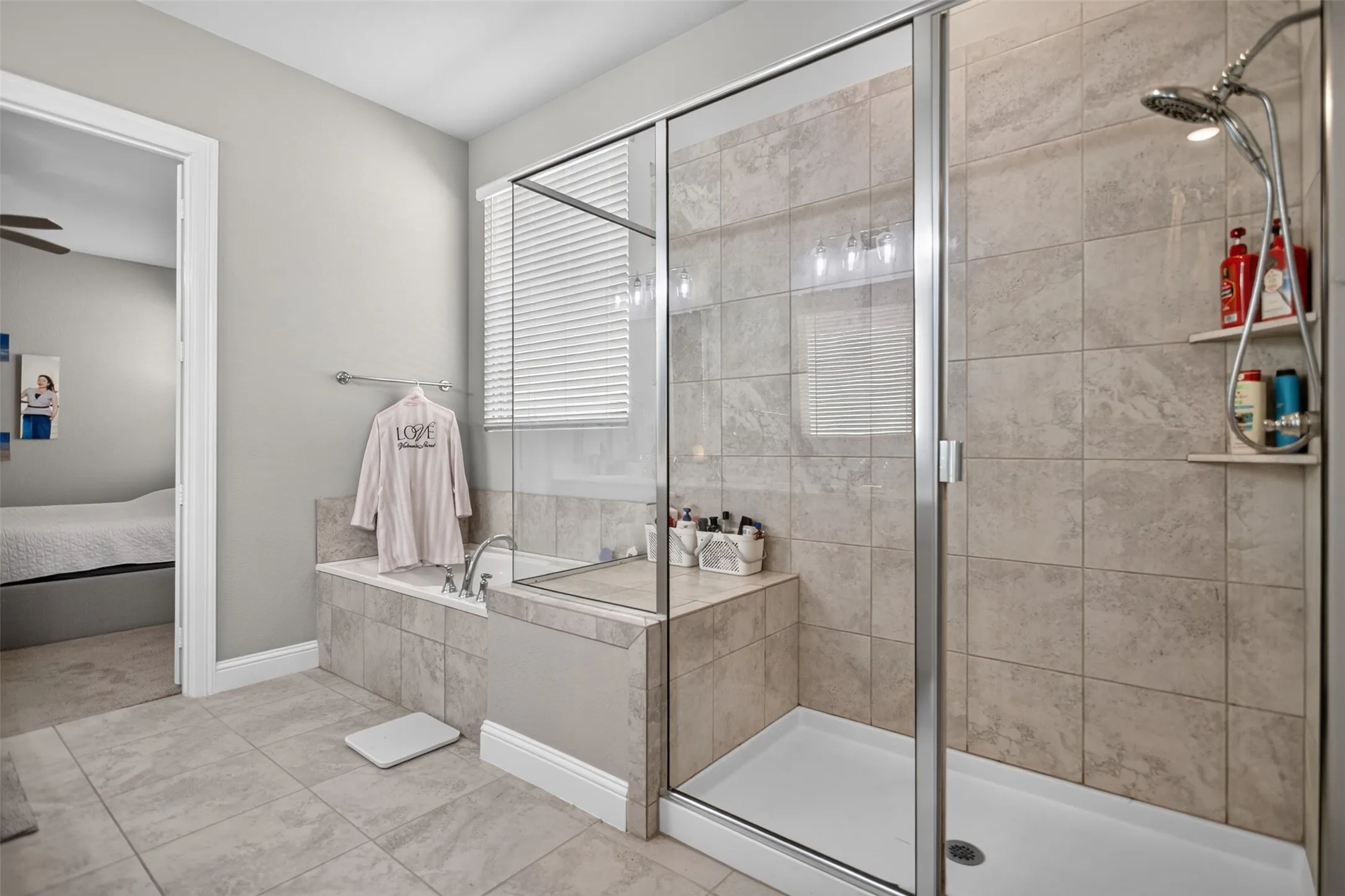 Tiled flooring, glass-enclosed shower with a built-in shelf, tiled shower surround, recessed bathtub, and window with blinds
