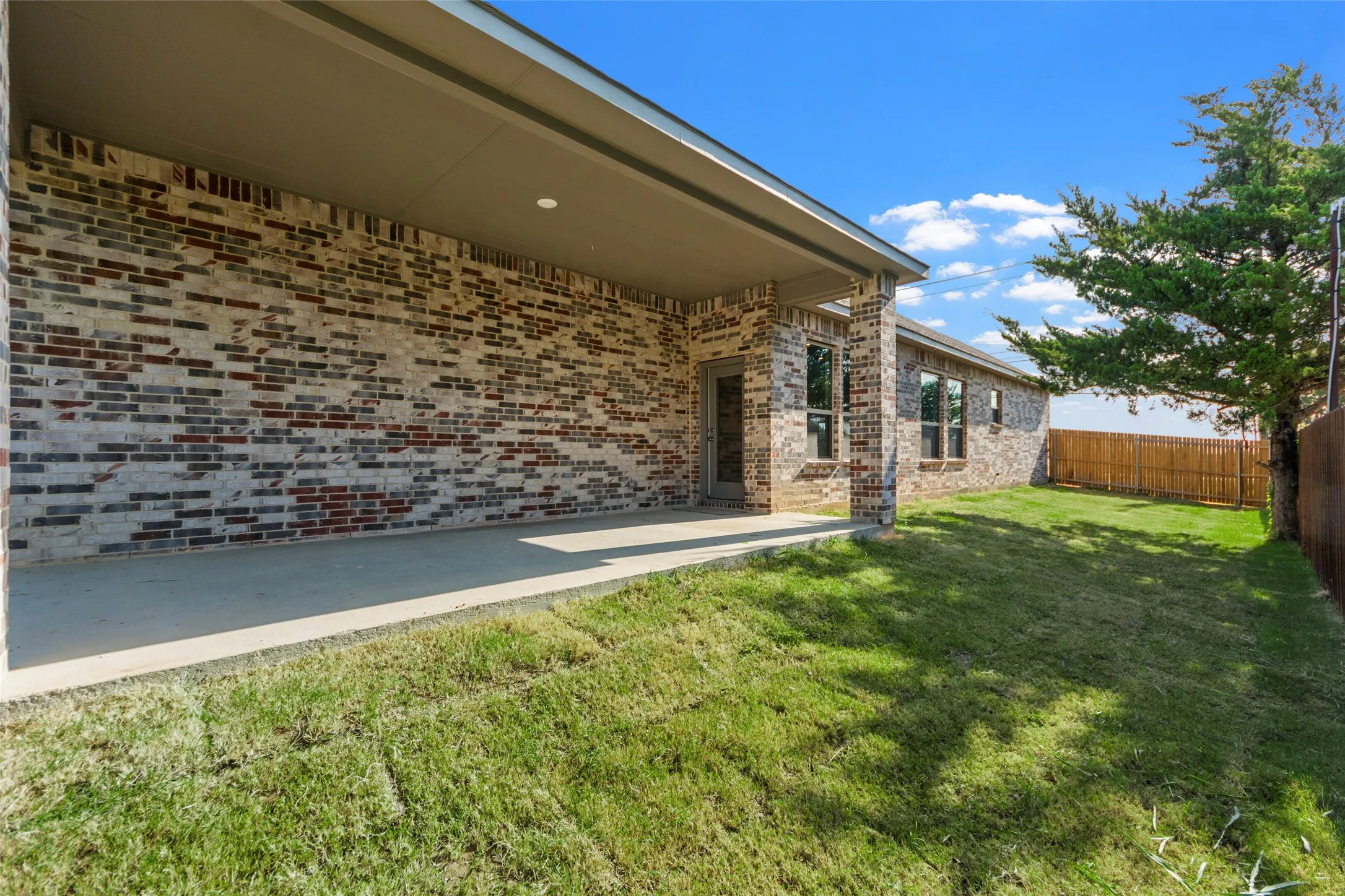 Covered concrete patio with recessed lighting, brick exterior, and a fenced yard with established trees
