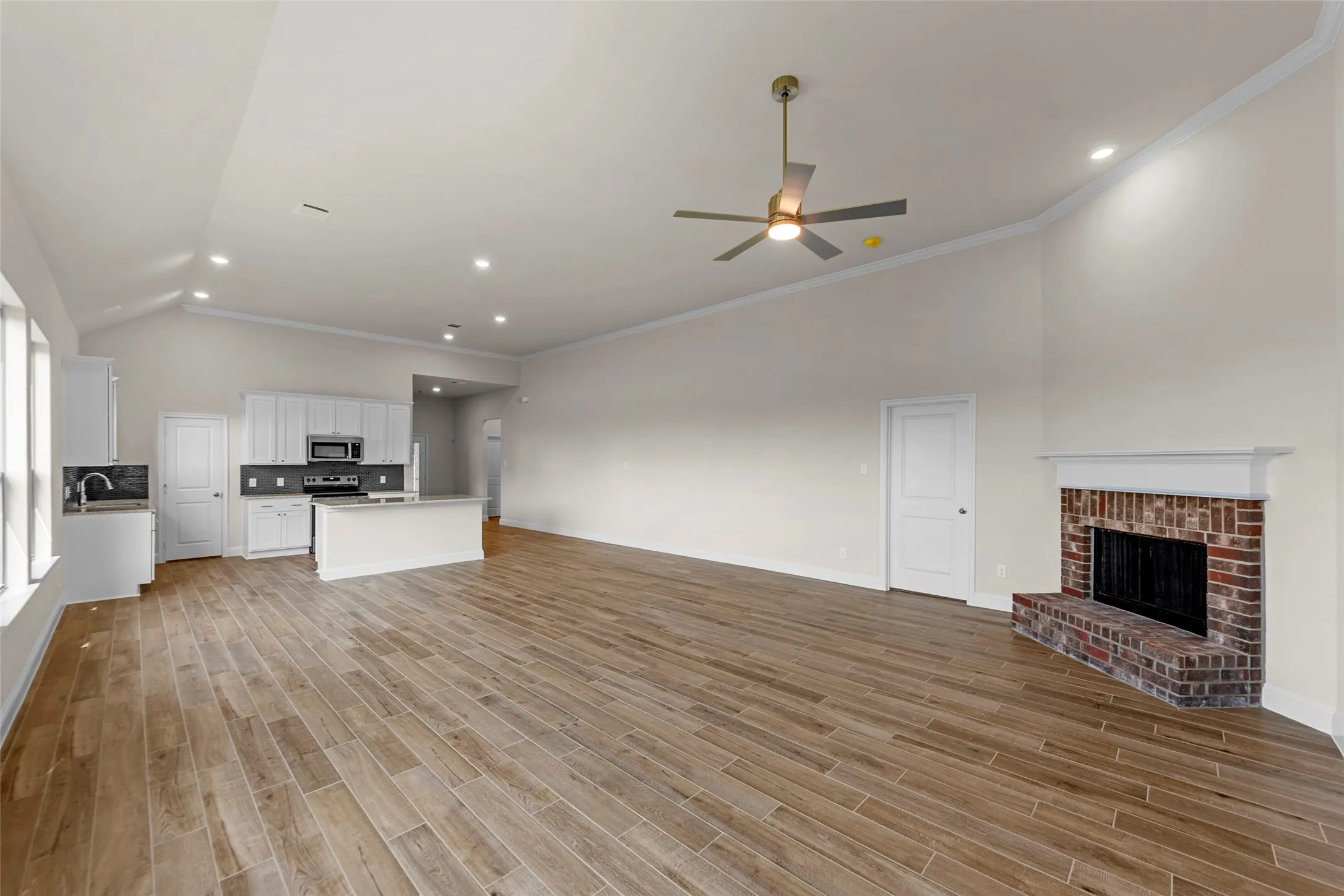 Spacious open-concept interior featuring vaulted ceilings, wood-finish flooring, a brick fireplace, and a kitchen with white cabinetry and stainless steel appliances