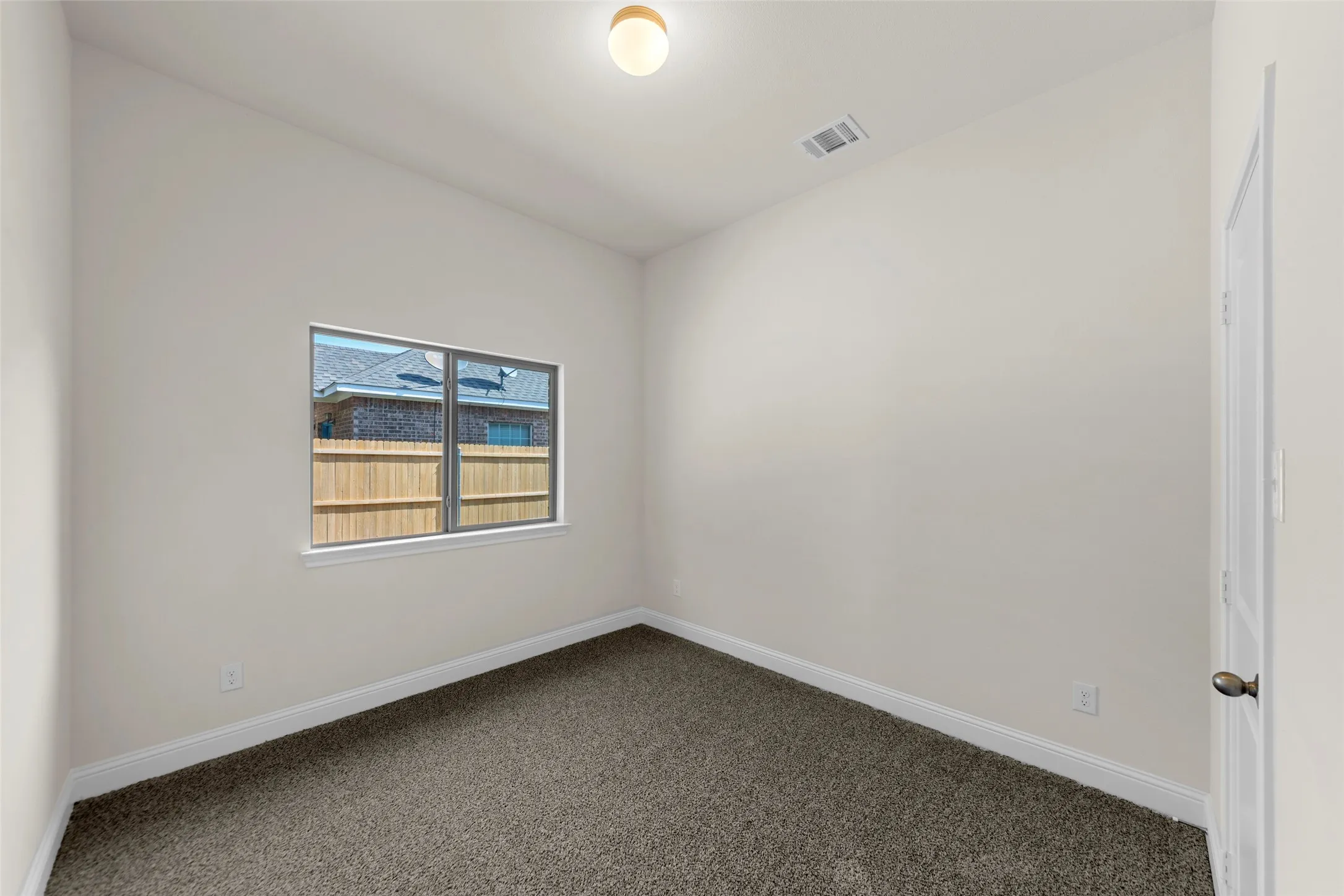 Neutral wall paint, textured carpet flooring, window with exterior fencing, white trim, and a ceiling-mounted light fixture