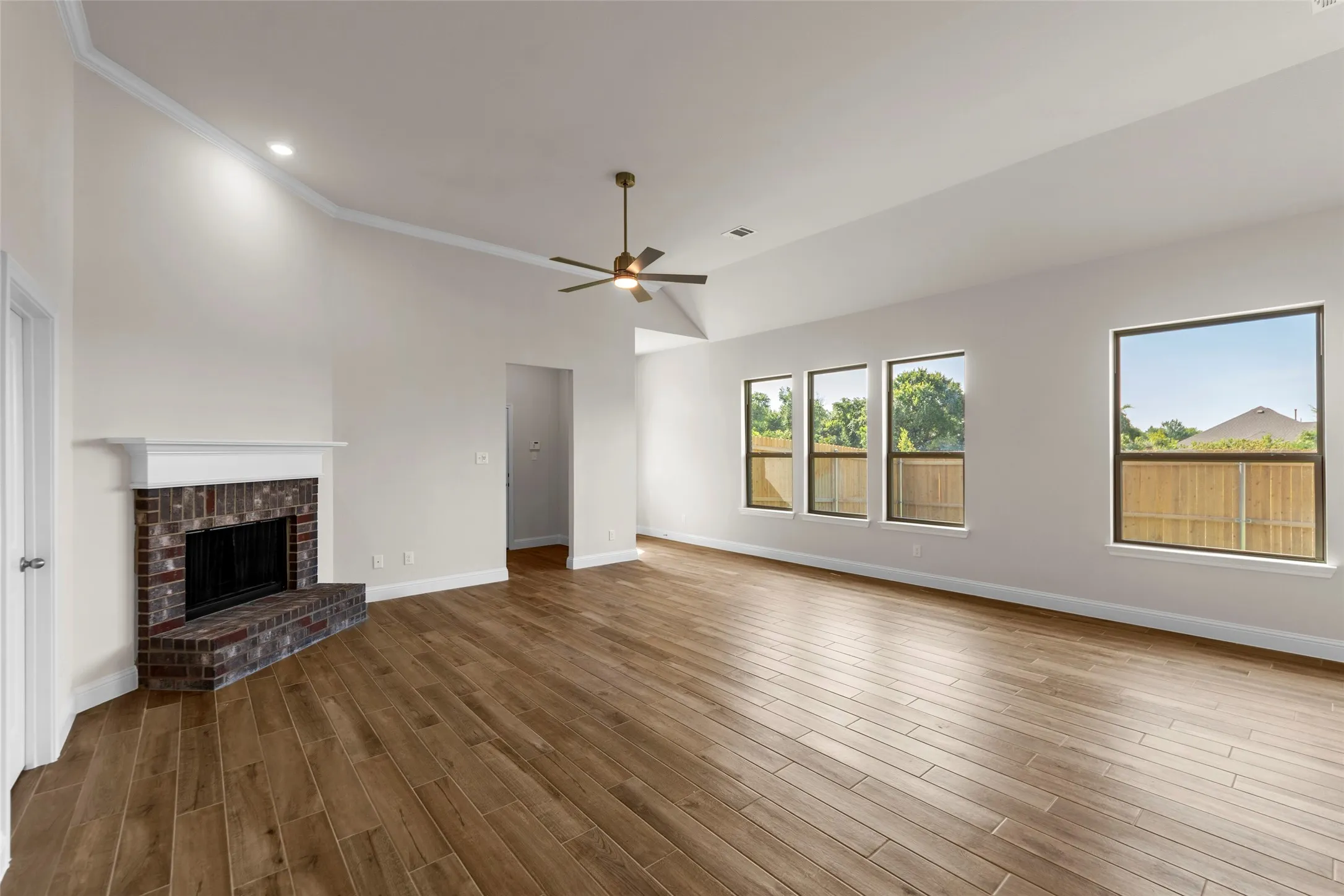 Vaulted ceilings with crown molding, brick fireplace with white mantel, wood-finish flooring, and multiple windows