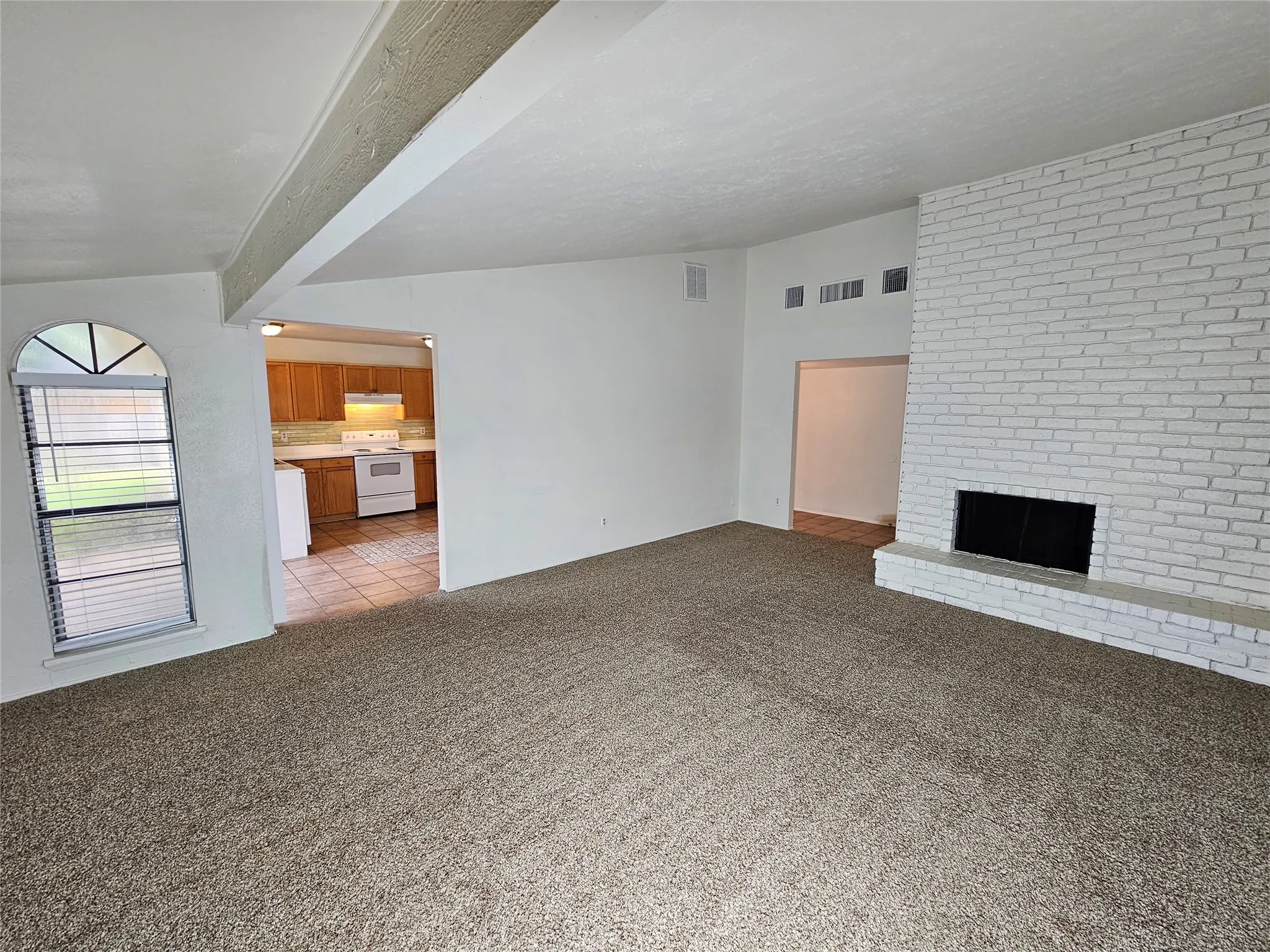 Spacious living area featuring a white-painted brick fireplace with hearth, textured beam ceiling accent, and neutral carpet flooring