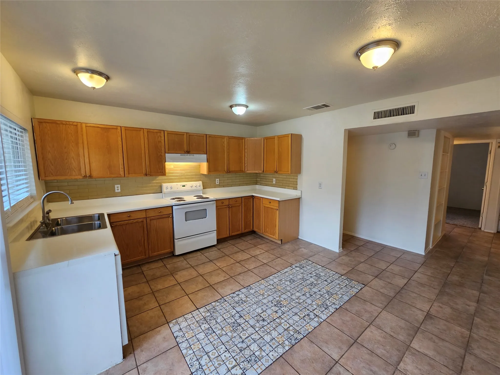 Kitchen featuring wood-finish cabinetry, a double basin stainless steel sink, white countertops, tile backsplash, and tile flooring