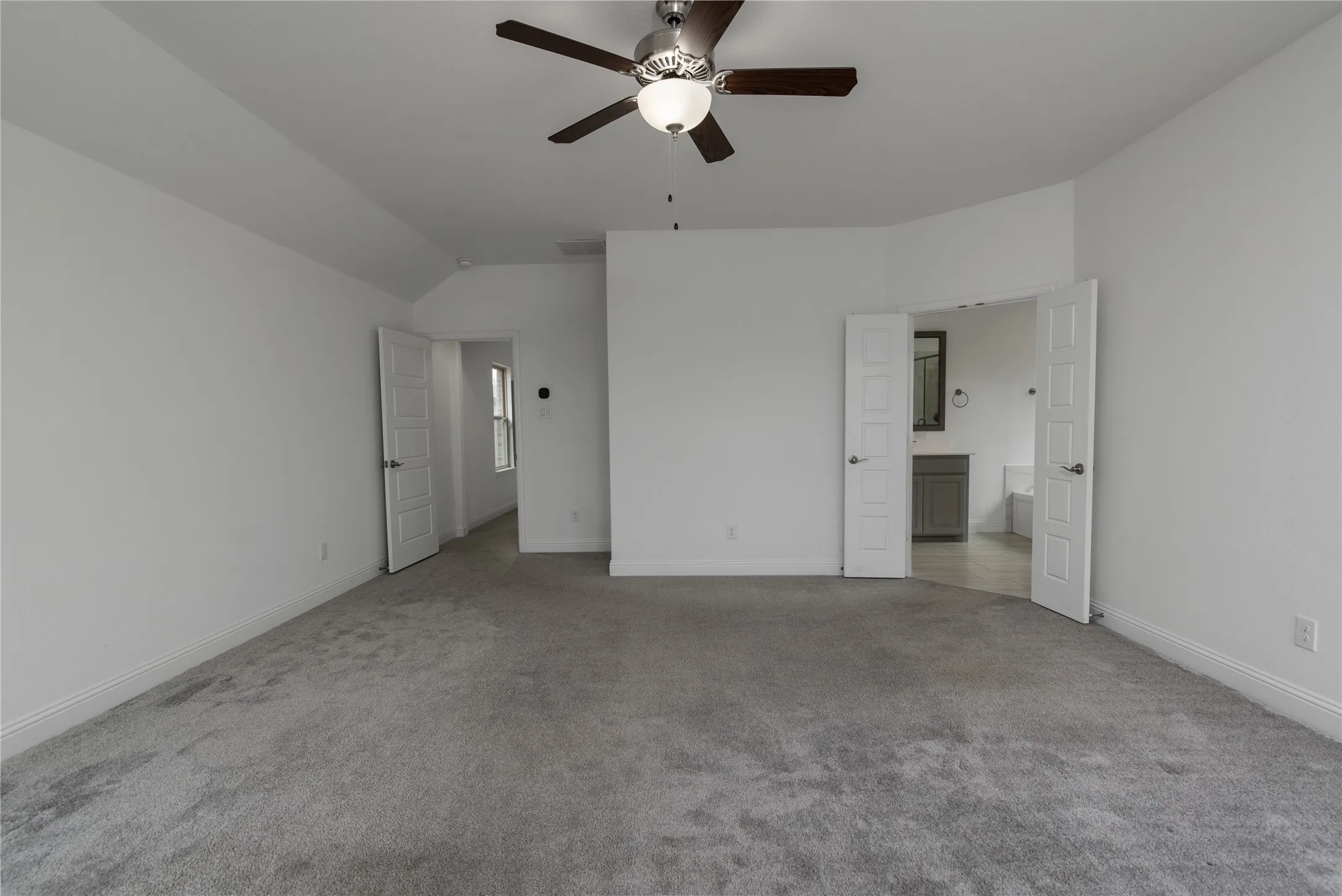 Spacious room with vaulted ceiling, ceiling fan, plush carpeting, and double-door entry to a bathroom featuring a vanity with a mirror and a soaking tub