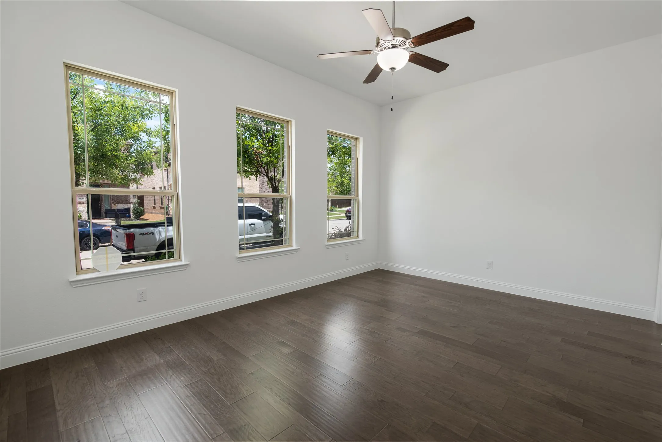 Spacious room featuring wood-finish flooring, three large windows, a ceiling fan with integrated lighting, and neutral wall tones