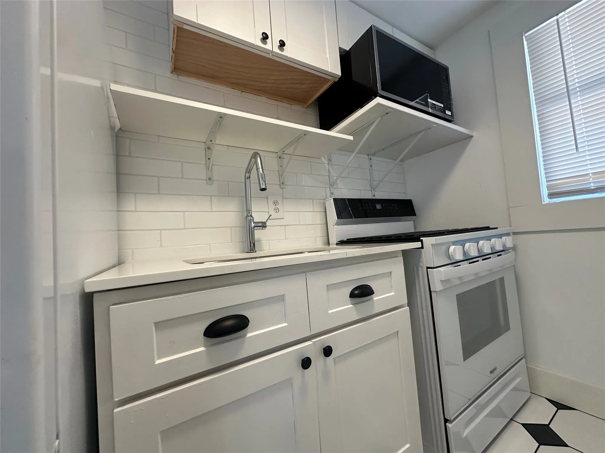 White shaker-style cabinetry with matte black hardware, subway tile backsplash, solid surface countertops, stainless steel sink with a gooseneck faucet, and geometric floor tiling