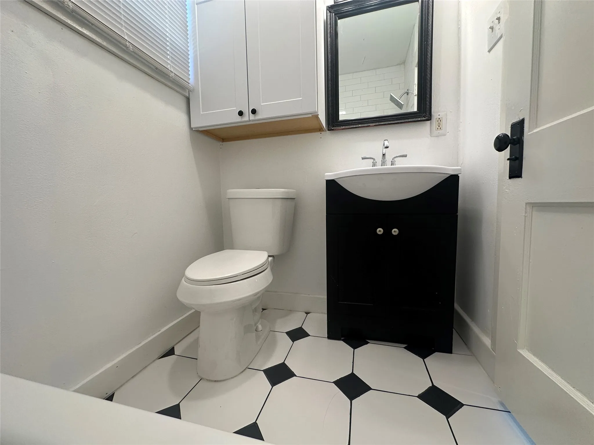 Bathroom featuring white walls, black and white hexagonal floor tiles, a white toilet, a black vanity with an integrated white sink, and a mirrored medicine cabinet